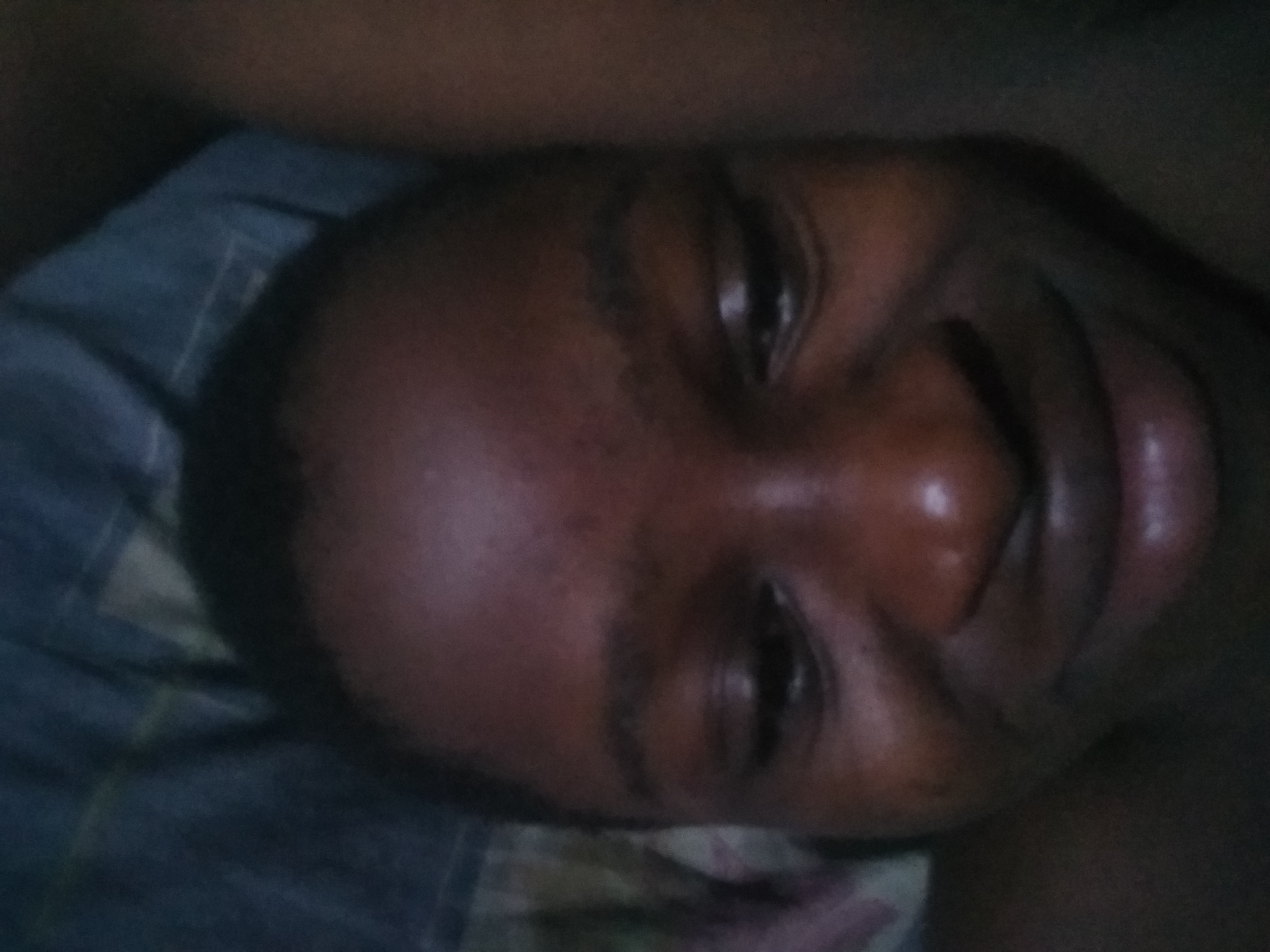 A close-up, slightly blurred photo shows a person lying on what appears to be a patterned, blue and gray fabric. The person has dark skin, dark hair, and a large nose. Their eyes are open. Due to the extreme close-up and lack of environmental context, it is impossible to provide details about Shangisha, Nigeria or any activities or interactions.