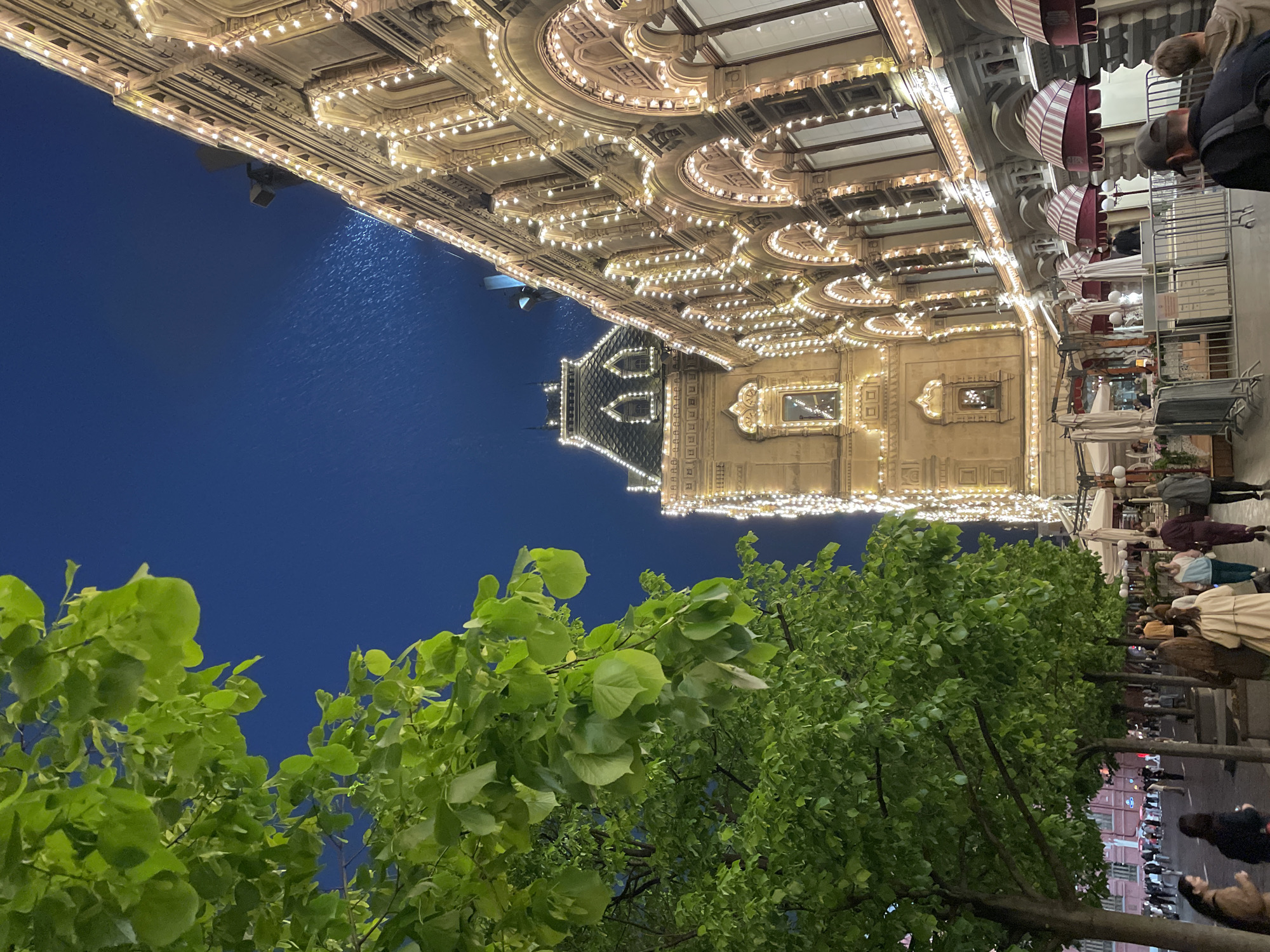 This is a nighttime photo of Moscow, Russia. The GUM department store is in the upper section of the image, lit up with many lights. There are people on the sidewalk alongside the store. In the foreground are leafy trees. The sky above is a dark blue.