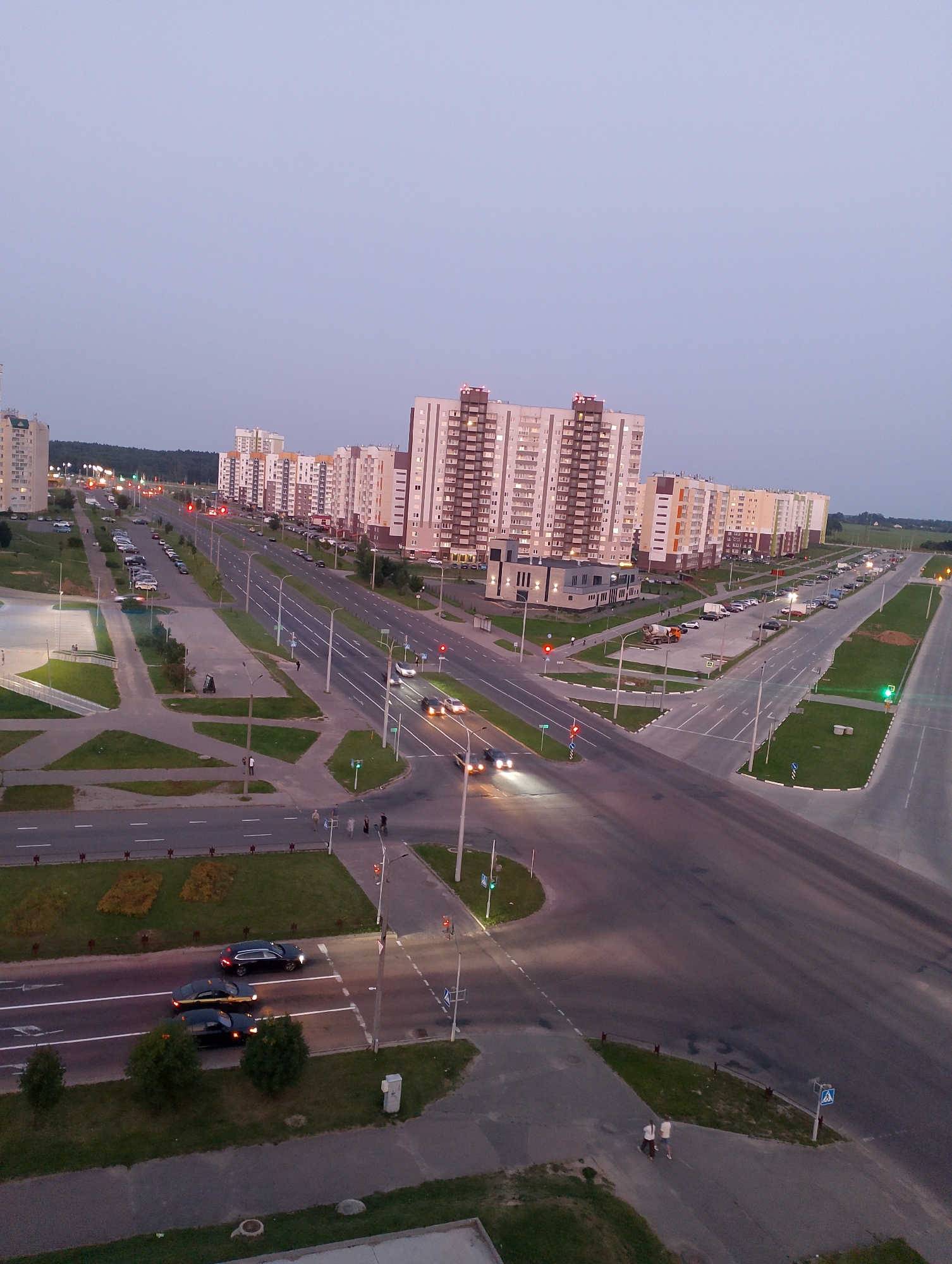 This is an aerial, high-angle shot of an intersection in Vitsebsk, Belarus, at dusk or dawn. A multi-lane road runs diagonally from the bottom left to the upper right, with another road intersecting it from the lower right. Several apartment buildings, ranging from 10 to approximately 15 stories, are visible in the background, with lights on in many windows. Traffic lights are present at the intersection, with some showing green and others red. Cars are driving on the roads, including several black sedans and a dark SUV. There are streetlights lining the roads. Pedestrians are visible walking on sidewalks near the intersection. Grass and some trees are present in the green spaces around the roads and buildings. The sky is a pale, overcast blue.