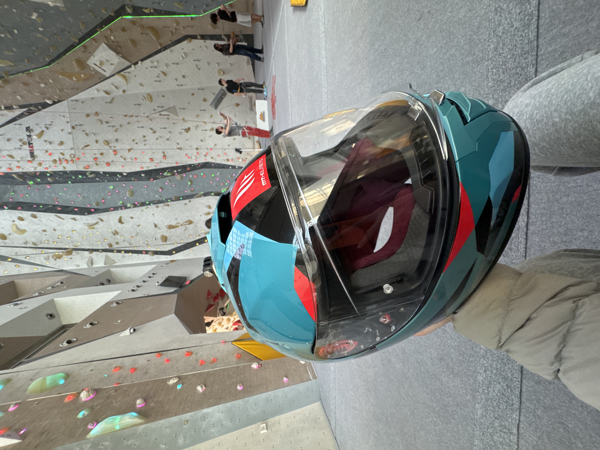 A close-up photograph shows a blue and black motorcycle helmet with a clear visor held in someone's hand. The background is slightly blurred but reveals an indoor rock climbing facility with various climbing holds on the walls. Several people can be seen in the background, some appearing to be actively climbing. The location is likely in Sofia, Bulgaria.