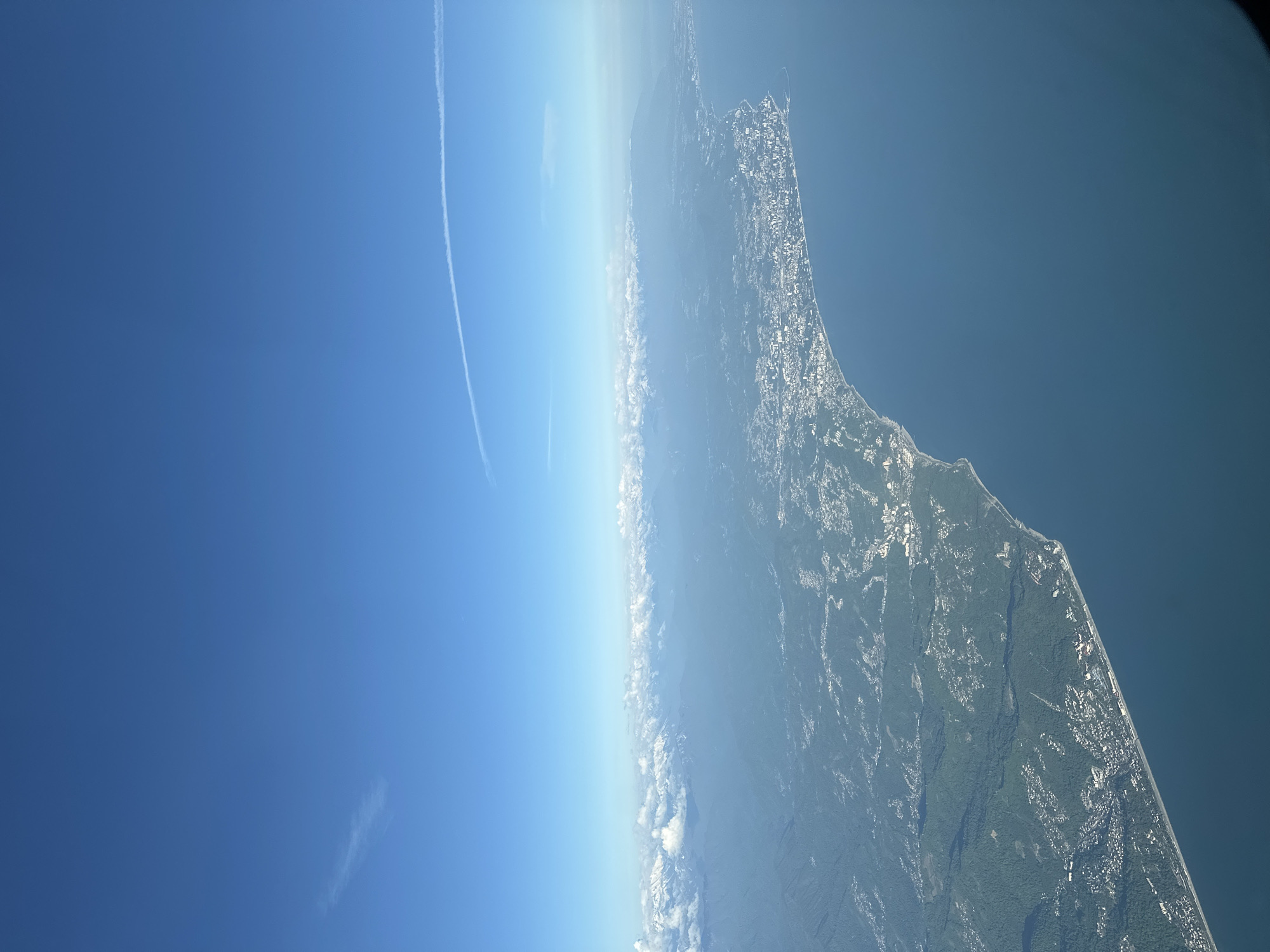The image shows a view from a high altitude, possibly from an airplane or a high vantage point, oriented vertically. The sky is clear blue on the left. The land is on the right, showing a shoreline with vegetation, possible structures, and an urban area. The clouds are visible along the horizon. It is impossible to locate Vishnevka, Russia in the image.