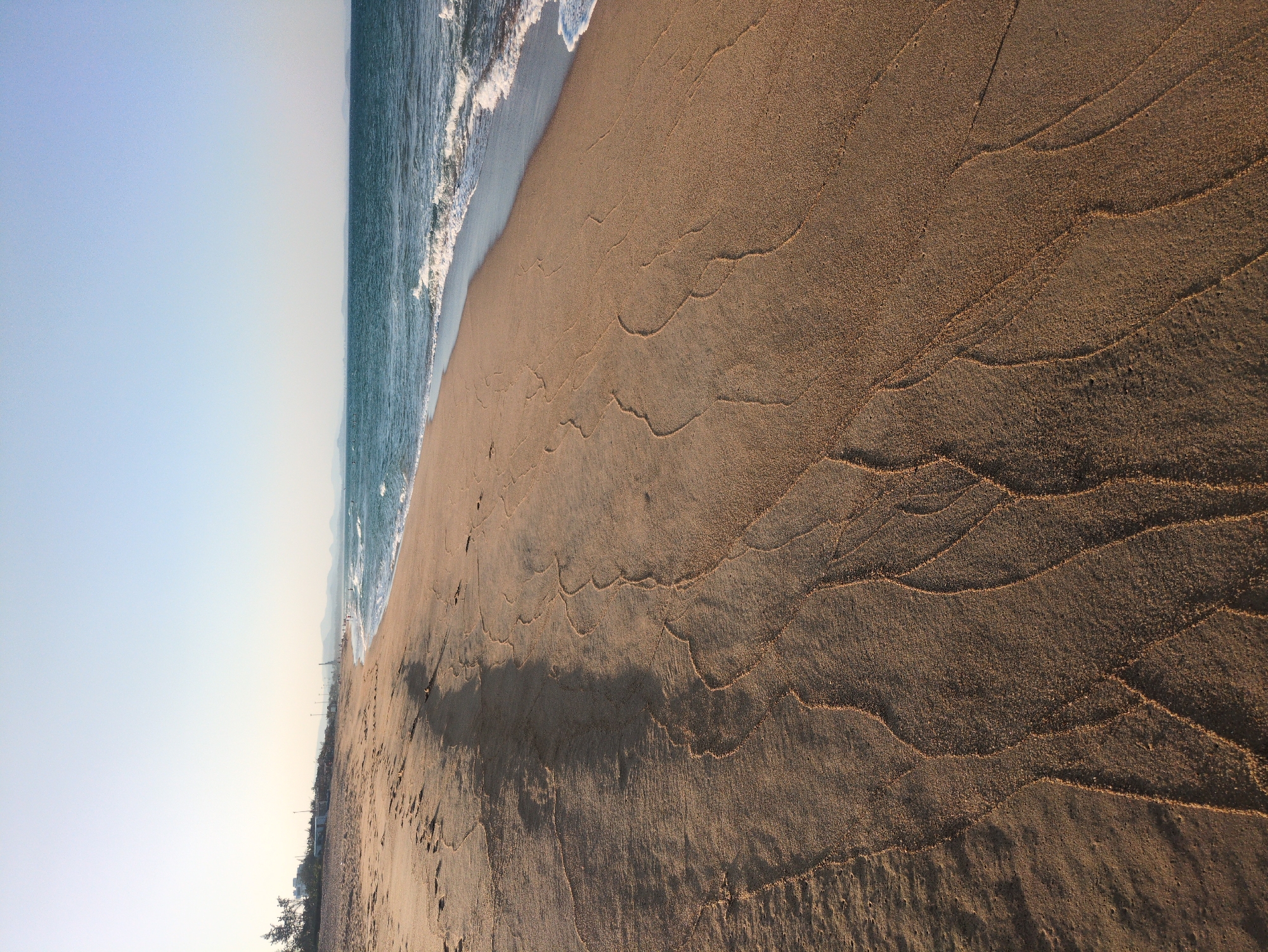 The image shows a beach in Tuy Hòa, Vietnam, with the ocean on the left side and the sandy beach on the right. The sky is clear and light blue. Waves are visible along the shoreline. The sand is marked with wavy patterns, possibly from the tide. A shadow stretches across the sand. A distant cityscape is seen at the edge of the beach.