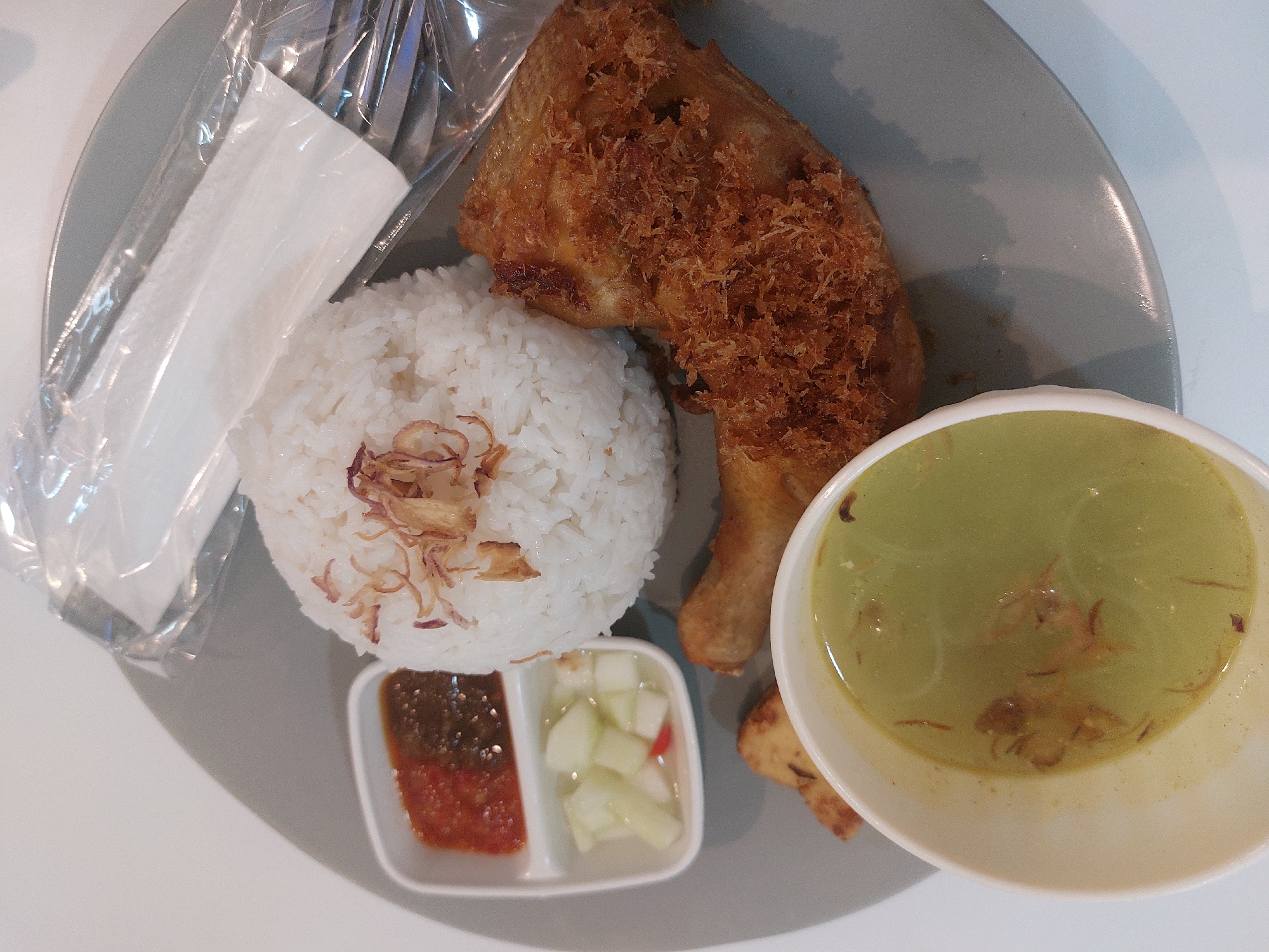 A plate of food, likely nasi ayam, is presented on a gray circular plate. The dish includes a generous portion of white rice, topped with fried shallots. To the left of the rice are two small white rectangular containers; the top one holds a reddish sauce, possibly sambal, and the bottom one contains diced cucumbers with a hint of red chili. A piece of fried chicken, with a crispy, golden-brown coating, is prominently featured on the right side of the plate. In the lower part of the image, a small white bowl contains a light green soup, possibly soto ayam, also garnished with fried shallots. A few pieces of fried potato are visible near the soup bowl. A clear plastic wrapper covers cutlery, including a fork and knife, resting on the upper right side of the plate. The setting appears to be an indoor dining area, and while the specific location within Nunukan, Indonesia, cannot be determined from the image alone, such a dish is commonly found in Indonesian eateries.
