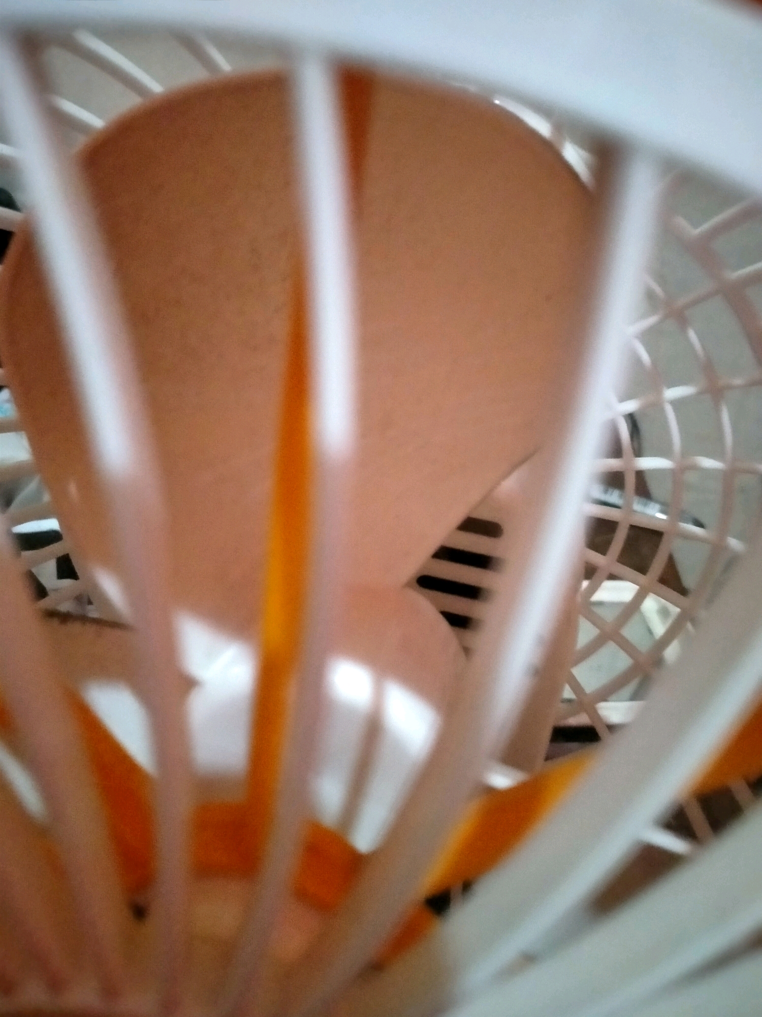 The image shows a close-up, out-of-focus view of a fan, likely in a domestic setting in Klepu, Indonesia. The fan has a white cage with vertical bars. Inside the cage, a light orange fan blade is visible, with some shadows and reflections. The background is blurred and appears to be a dark, patterned surface. No people or specific actions are discernible. The overall impression is of a still object, viewed from a close proximity.