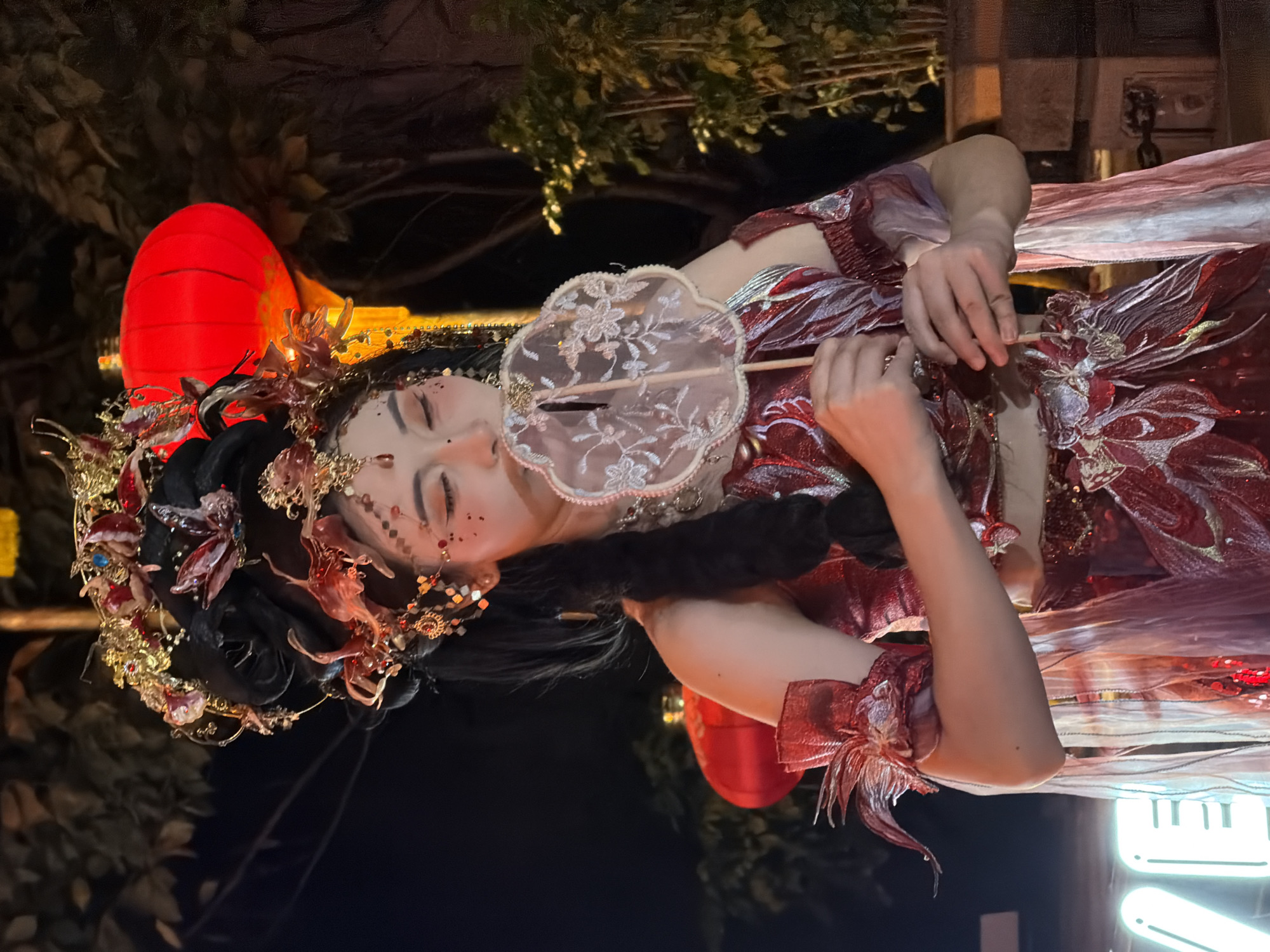 In Yuzhong District, China, a woman in a detailed, red and gold traditional Chinese costume with elaborate hair ornaments and makeup poses at night. She is holding a small, ornate fan to her face. Red lanterns are visible in the background, along with trees and a neon sign partially showing the letters "VE". The woman is positioned centrally in the frame, with her body angled slightly to the right. The lighting suggests an outdoor evening event or performance.