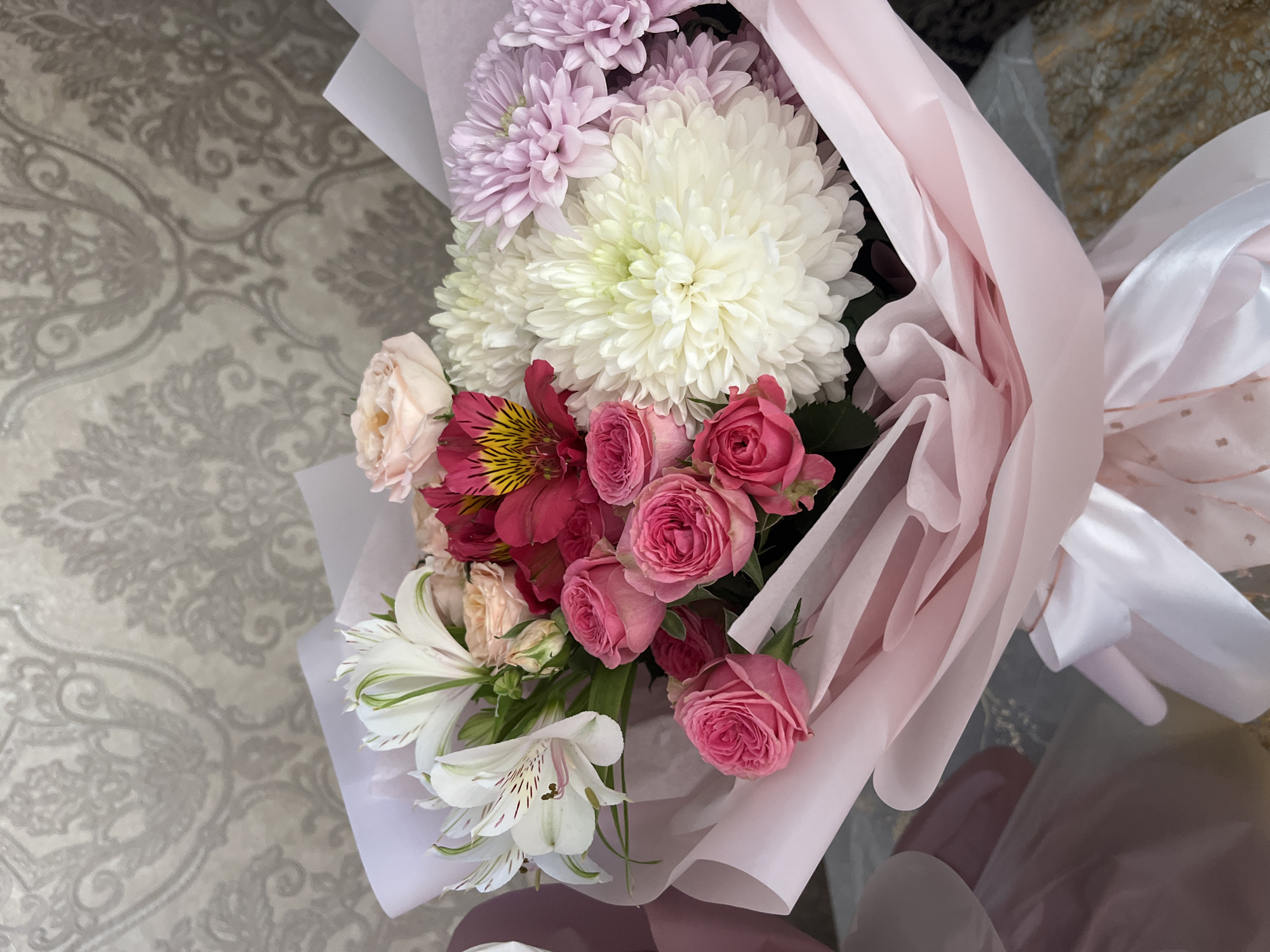 The image shows a bouquet of flowers wrapped in pink tissue paper. The bouquet contains white chrysanthemums, light purple asters, pink roses, peach-colored roses, and white alstroemeria with red and yellow accents. The bouquet is lying on a surface with a patterned design. The location in the city of Kaspiysk, Russia is not evident from the image.