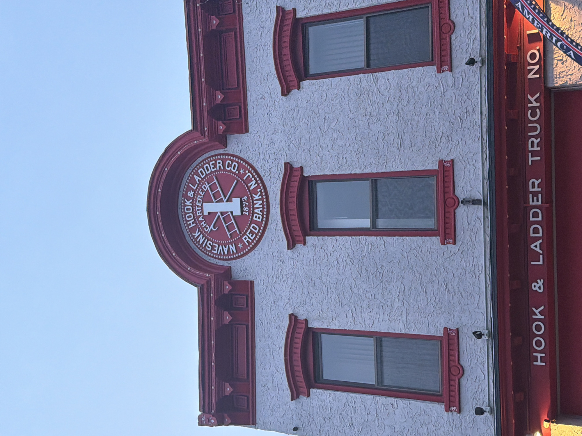This is a waist-up, eye-level, outdoor, daylight photograph of the front of the historic Navesink Hook & Ladder Co. building in Red Bank, New Jersey, United States of America. The building is painted white with red trim. The most prominent feature is a circular emblem at the top of the building that reads "NAVESINK HOOK & LADDER CO. CHARTERED 1873 RED BANK, N.J.". Below the emblem are three rectangular windows with red trim and arched red cornices. The facade is textured stucco. At the bottom of the image, a red structure with white lettering reads "HOOK & LADDER TRUCK NO. 1", indicating it is a firehouse. An American flag is partially visible on the right side of the frame. The sky is clear and blue. There are no people or vehicles visible in the image.