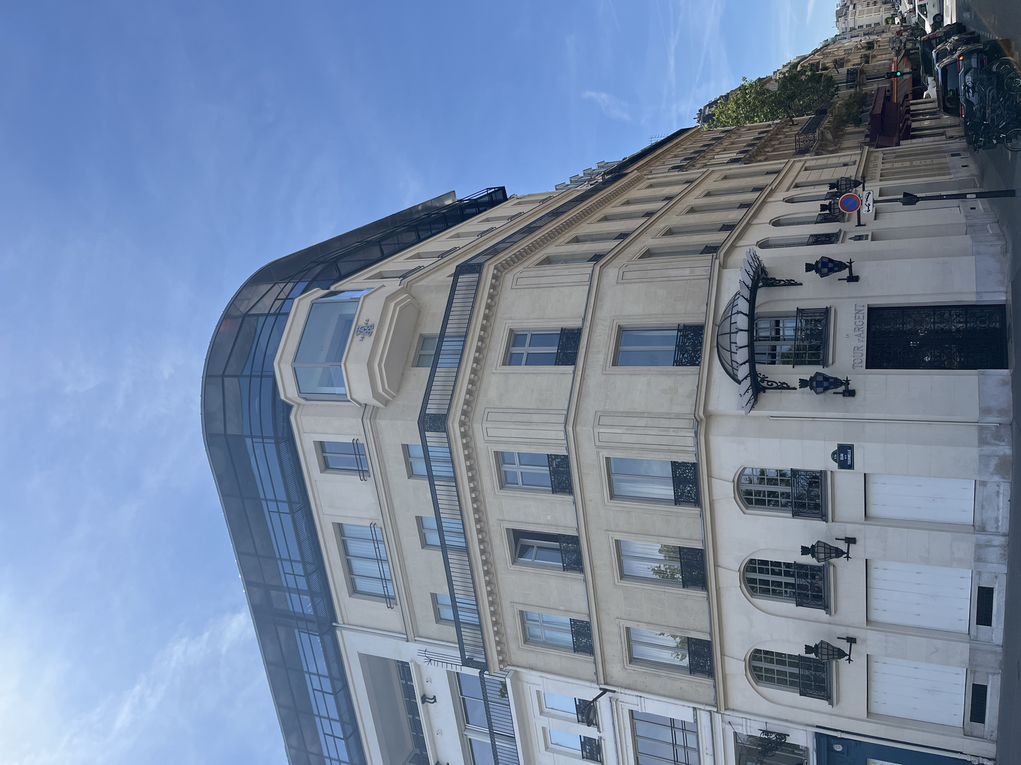 The image shows the facade of a building in Paris, France, on a sunny day. The building is tall and appears to be in an urban setting, with other buildings visible on the street. The building itself has multiple stories, with a combination of stone and modern glass architecture. The lower floors feature arched windows and decorative lamps, with white shuttered doors. Above, there are rows of rectangular windows with balconies and ornate iron railings. The top of the building is a modern, curved glass structure. A street sign reading "QUA TOWERNELL' is visible on the building. Cars and bicycles are parked on the street in front of the building. The sky is a clear blue with a few wispy clouds.