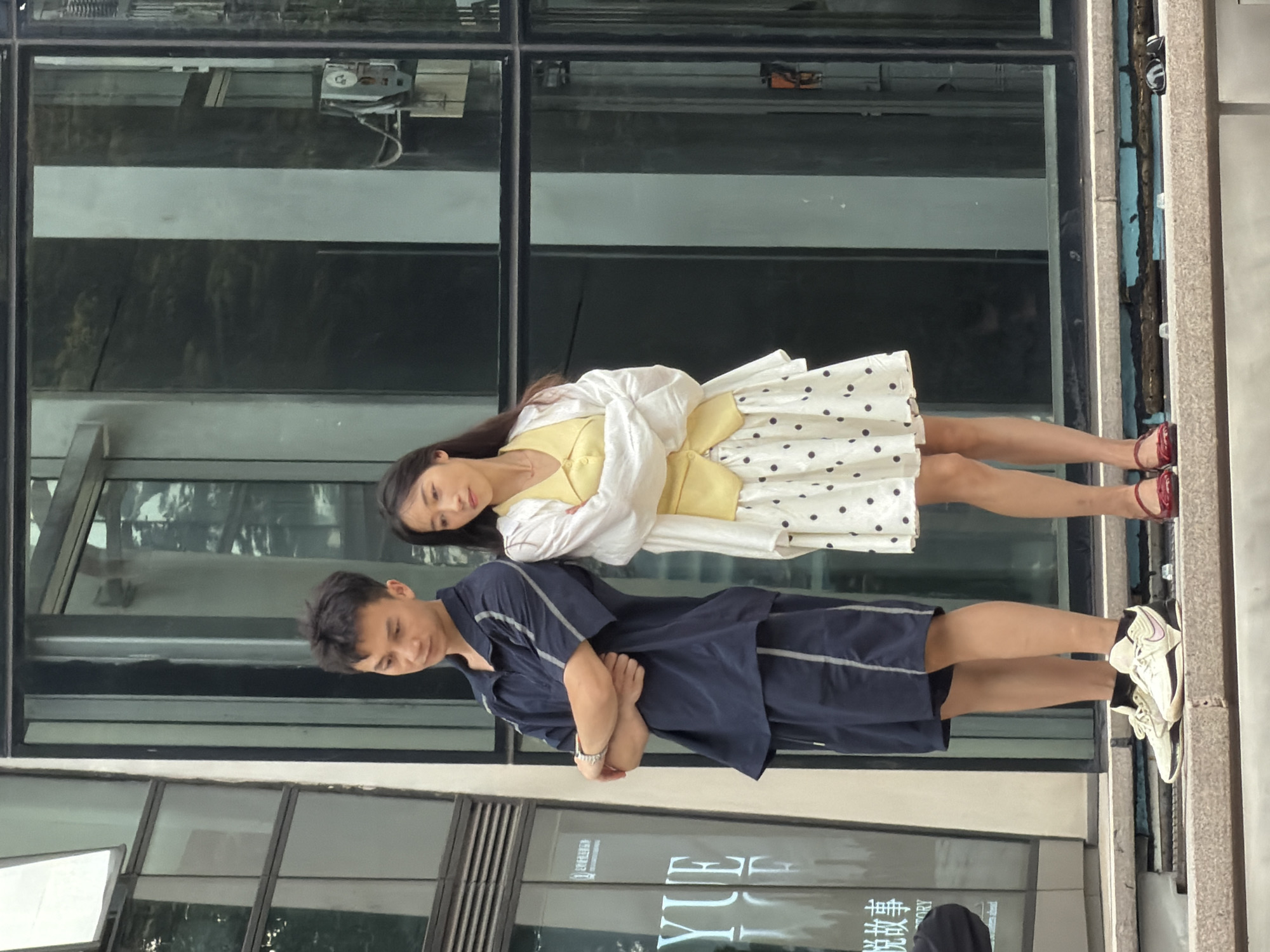In Yuzhong District, China, a man and a woman stand outdoors with their backs to each other. The man, on the left, wears a dark blue short-sleeved shirt with gray stripes and matching shorts. He has his arms crossed and wears white and black sneakers. The woman, on the right, is dressed in a yellow top, a white cardigan, and a white skirt with black polka dots. She wears red flat shoes. They are standing on a ledge in front of a building with large, dark windows.