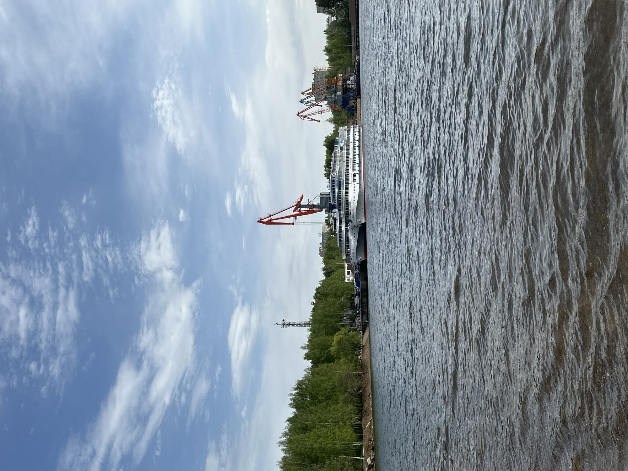 A body of water occupies the right side of the frame, its surface characterized by ripples and a slightly gray tone. Adjacent to the water is a strip of land covered in trees, forming a green barrier. Further beyond the trees, an assortment of cranes stands in the distance. A tall antenna tower can be seen near the trees. In the top center is a white, multi-story passenger vessel, presumably docked. Above the scene is a sky filled with blue and scattered clouds.