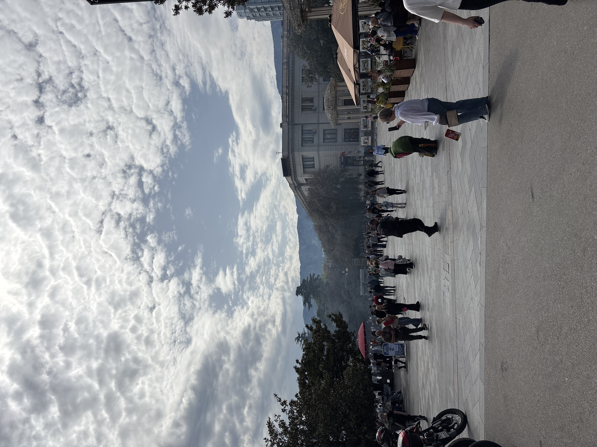 The image shows a busy city street scene. A crowd of people is walking on a paved pedestrian area. Buildings line the street on one side, with mountains visible in the background. The sky is filled with clouds. A cafe with outdoor seating is present. A motorcycle is parked on the right side of the image. People are interacting with each other and some are looking at their phones. The location is город Ялта, Ukraine.