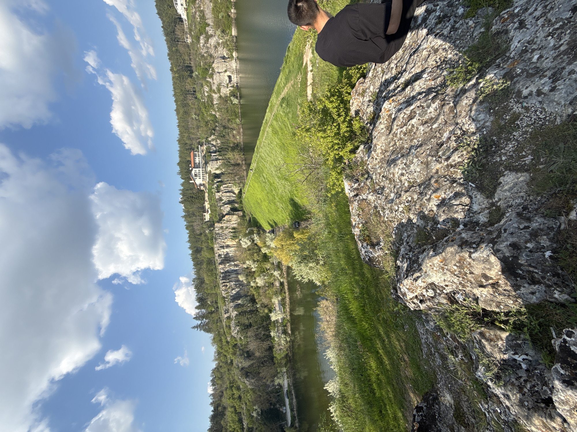 The image presents a scenic vista from a high vantage point, likely a cliff edge, overlooking a natural landscape in Pleven, Bulgaria. The scene is bathed in bright daylight under a clear blue sky dotted with scattered white cumulus clouds.
In the immediate foreground, a person, seen from behind and wearing a black t-shirt, is seated or crouching on a rugged, lichen-covered rock outcrop, looking out over the view. The rocky terrain, with its varied textures and patches of green growth, dominates the right side of the foreground.
Below and extending into the mid-ground, a vibrant green grassy area or field slopes gently towards a calm body of water, which appears to be a river or lake. The water's surface reflects the sky and surrounding greenery, appearing dark green or olive.
On the far side of the water, the landscape rises into steep, rocky cliffs and hillsides densely covered with trees and lush vegetation, forming a verdant backdrop. Several multi-story buildings or houses with distinctive roofs, primarily white or light-colored, are visible nestled among the trees on the distant bank, suggesting human presence integrated into the natural environment.
The strong sunlight indicates a clear day, likely late morning or early afternoon, casting defined shadows across the landscape. The overall impression is one of peaceful contemplation amidst beautiful natural surroundings. No discernible text is visible in the image.