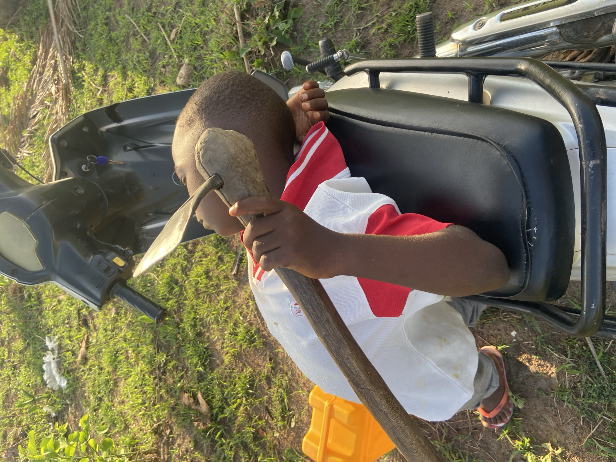 A child lies on their side on the ground near a fallen motorcycle. The location is Mararaba, Nigeria, based on the available information. The child wears a white shirt with red sleeves and gray pants. They are holding a gardening hoe. The motorcycle is silver and black with a black seat. The ground is covered in green grass. A yellow object is also visible near the child. The motorcycle has a key in the ignition.