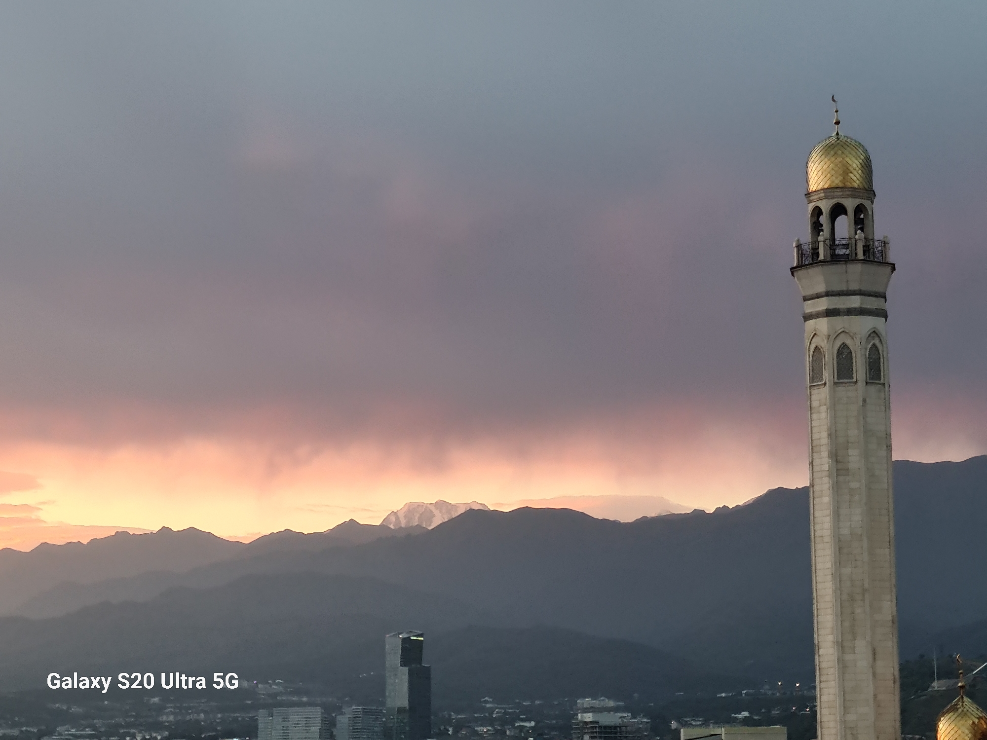 The image captures a cityscape at dusk, dominated by a towering minaret on the right side of the frame, topped with a golden dome and a crescent moon. In the background, a range of mountains stretches across the horizon, with their peaks catching the fading light of the sunset, tinged with pink and orange hues. The sky is a gradient of soft purples and grays, with clouds adding texture.
In the mid-ground, the silhouette of Almaty is visible, featuring a modern skyscraper and other buildings. The text "Galaxy S20 Ultra 5G" is overlaid in the bottom left corner, indicating the device used to capture the photograph. The overall scene depicts a serene urban landscape with a prominent religious structure against a dramatic natural backdrop.
