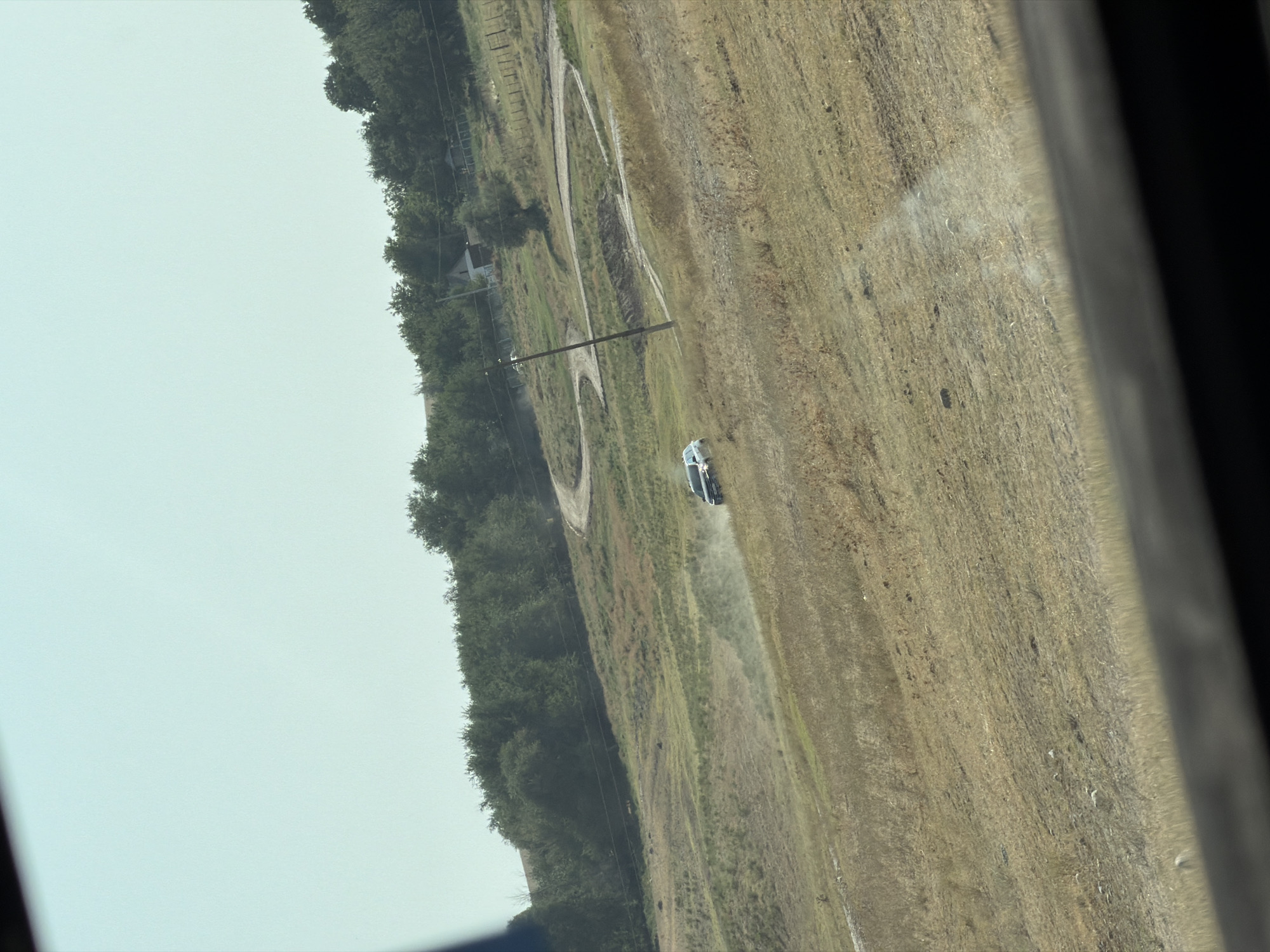 A car is driving on a winding dirt road that cuts through a dry, grassy hillside in a rural area. The car is kicking up a dust cloud as it moves. In the background, there are trees and some small buildings, suggesting a scattered rural settlement. The sky is clear and light blue. The scene appears to be captured from inside a vehicle, looking out through a window, as evidenced by the dark frame at the bottom of the image. The location is described as Теректі, Kazakhstan, indicating a place with natural, undeveloped terrain.