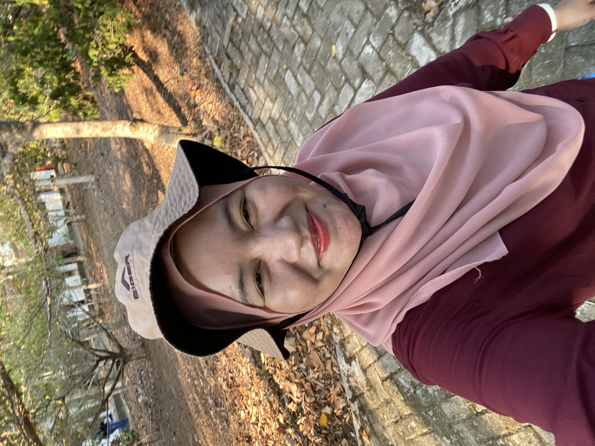 A woman wearing a maroon long-sleeved shirt, a pink hijab, and a beige bucket hat is taking a selfie outdoors in Sungai Pitung, Indonesia. She has fair skin, red lipstick, and is smiling. The background shows a brick pathway, dry leaves on the ground, trees, and a building. The sun is casting long shadows, indicating it is either early morning or late afternoon. A person in blue jeans and a dark shirt is visible in the far left background. The scene suggests a casual outdoor setting, possibly a park or a residential area.