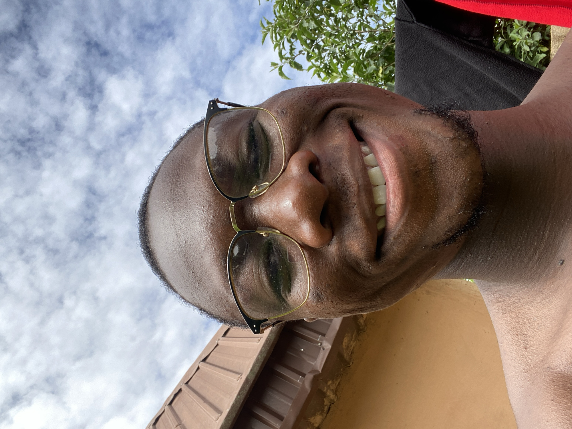 The image shows a close-up, wide-angle selfie of a Black man wearing glasses and smiling. He has a light beard and is outdoors. In the background, there is a brown roof and green foliage under a cloudy sky. The scene is likely a residential area in Bukuru, Nigeria, given the context provided.