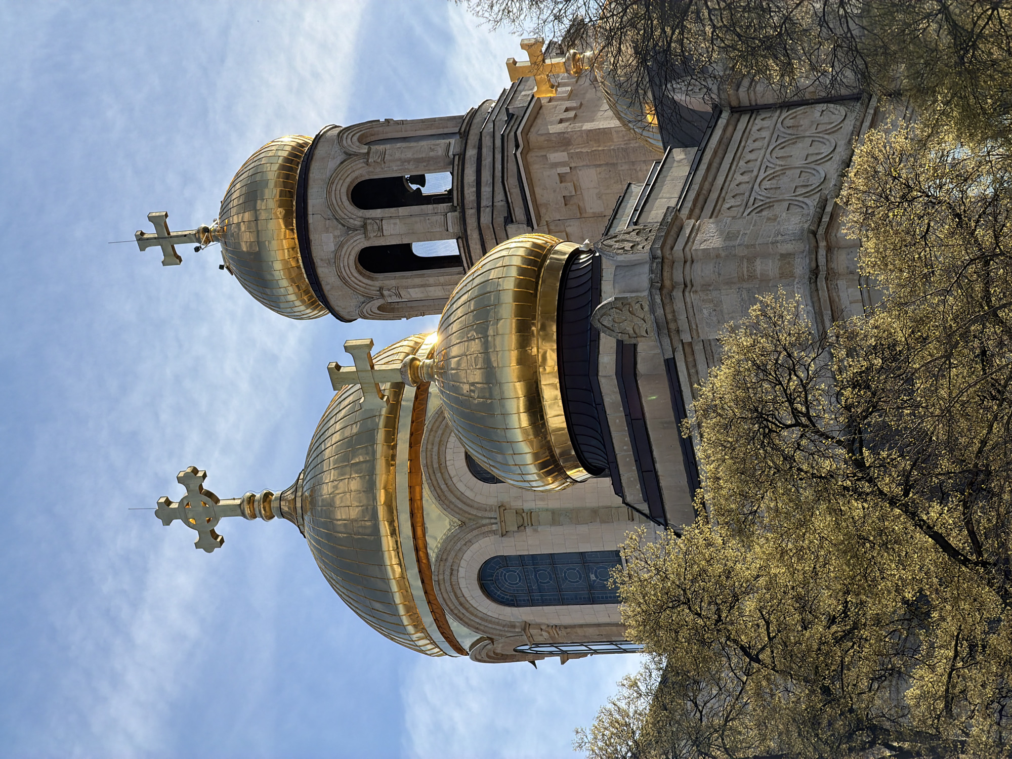 This media file presents a low-angle perspective of a grand Eastern Orthodox cathedral, likely the Dormition of the Theotokos Cathedral in Varna, Bulgaria. The central focus is on two shimmering golden domes, each topped with an ornate golden cross. The larger, more prominent dome is covered in reflective gold panels. To its right, a stone bell tower features a smaller golden dome and cross, along with arched belfry openings.
The cathedral's facade is constructed from light-colored stone, showcasing intricate architectural details. These include arched windows with dark, patterned glass, and sections of wall adorned with circular carved motifs, some enclosing cross shapes. The building is partially framed by tree branches with numerous small, light green leaves, suggesting early spring foliage. The background features a bright blue sky with scattered white clouds, indicating clear, daytime conditions. The image conveys the impressive scale and ornate beauty of the religious edifice. No text is visible within the image.