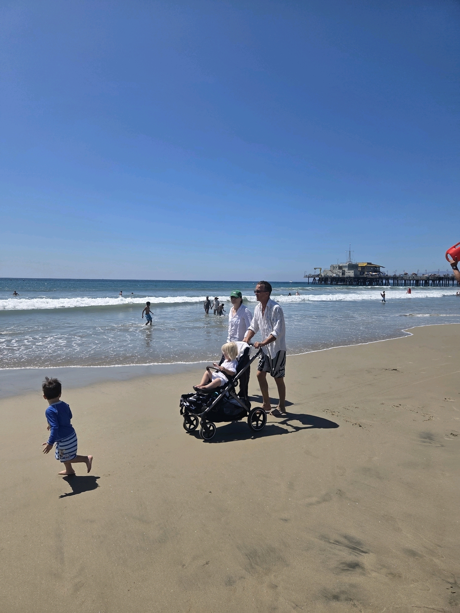 The image captures a sunny day at the Santa Monica Pier beach in Santa Monica, California. In the foreground, a young boy in a blue and white striped rash guard and swim trunks runs along the sand, casting a long shadow. To the right, a couple pushes a stroller with a young child in it. The man is wearing a white short-sleeved shirt and patterned shorts, while the woman wears a white top and a green cap. Behind them, several other people are visible, some wading in the ocean and others standing on the shore. The Santa Monica Pier, a prominent landmark, extends into the ocean in the background. The ocean is calm with gentle waves, and the sky is a clear, bright blue.