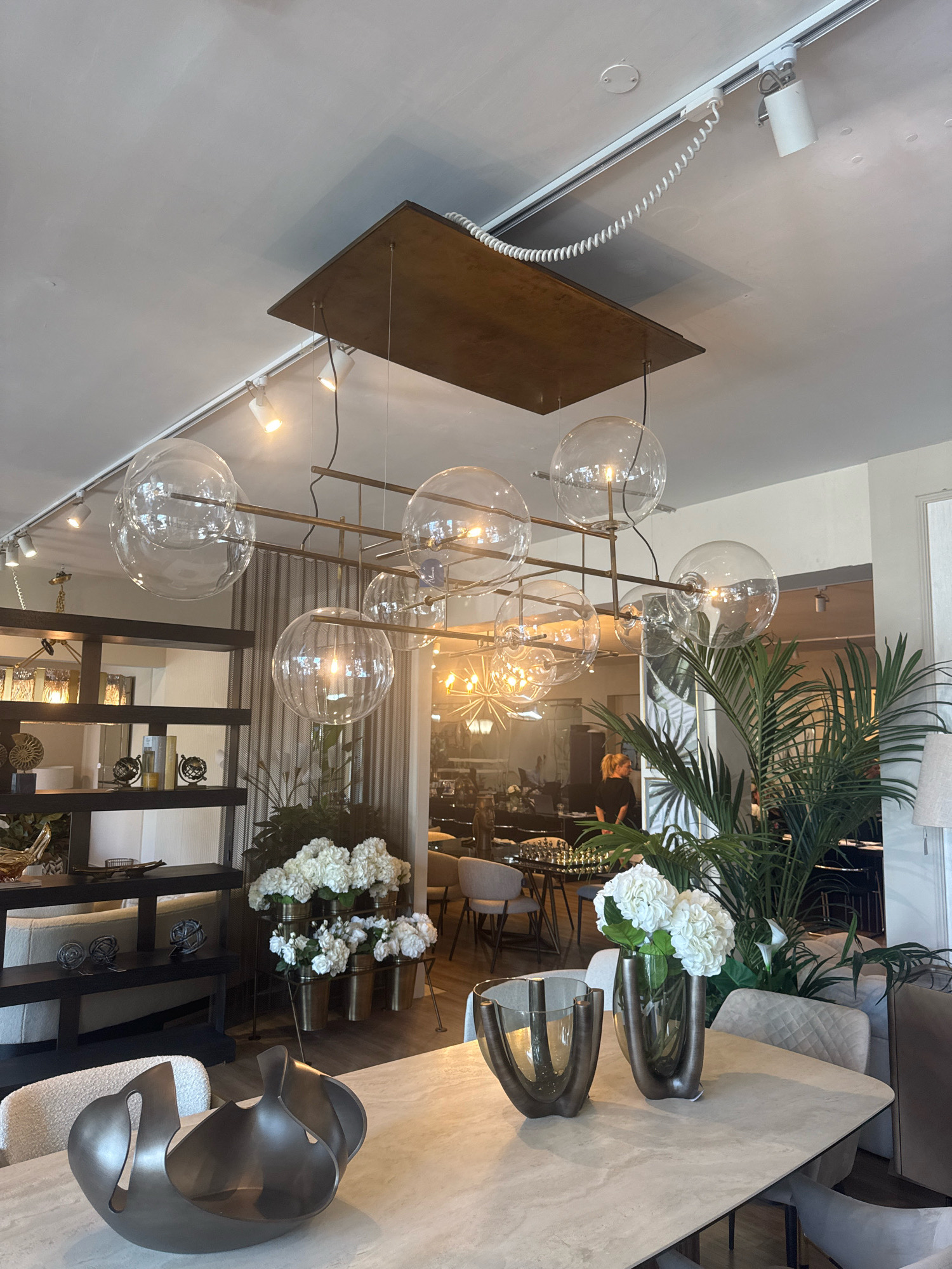 This is a photograph of the interior of a modern showroom or design studio in Agios Athanasios Municipality, Cyprus. The foreground features a light-colored dining table with two decorative vases and a sculptural centerpiece. One vase holds white flowers, while the other is empty. The centerpiece is a dark, metallic, abstract sculpture with organic curves. In the background, a large palm plant is visible, as are shelves filled with decorative items. A large, multi-globe chandelier with clear glass shades hangs from the ceiling, illuminated by spotlights. In the far background, several dining tables with chairs are set up, and a person is standing near a bar area. The overall aesthetic is contemporary and luxurious, with a focus on design elements and lighting.