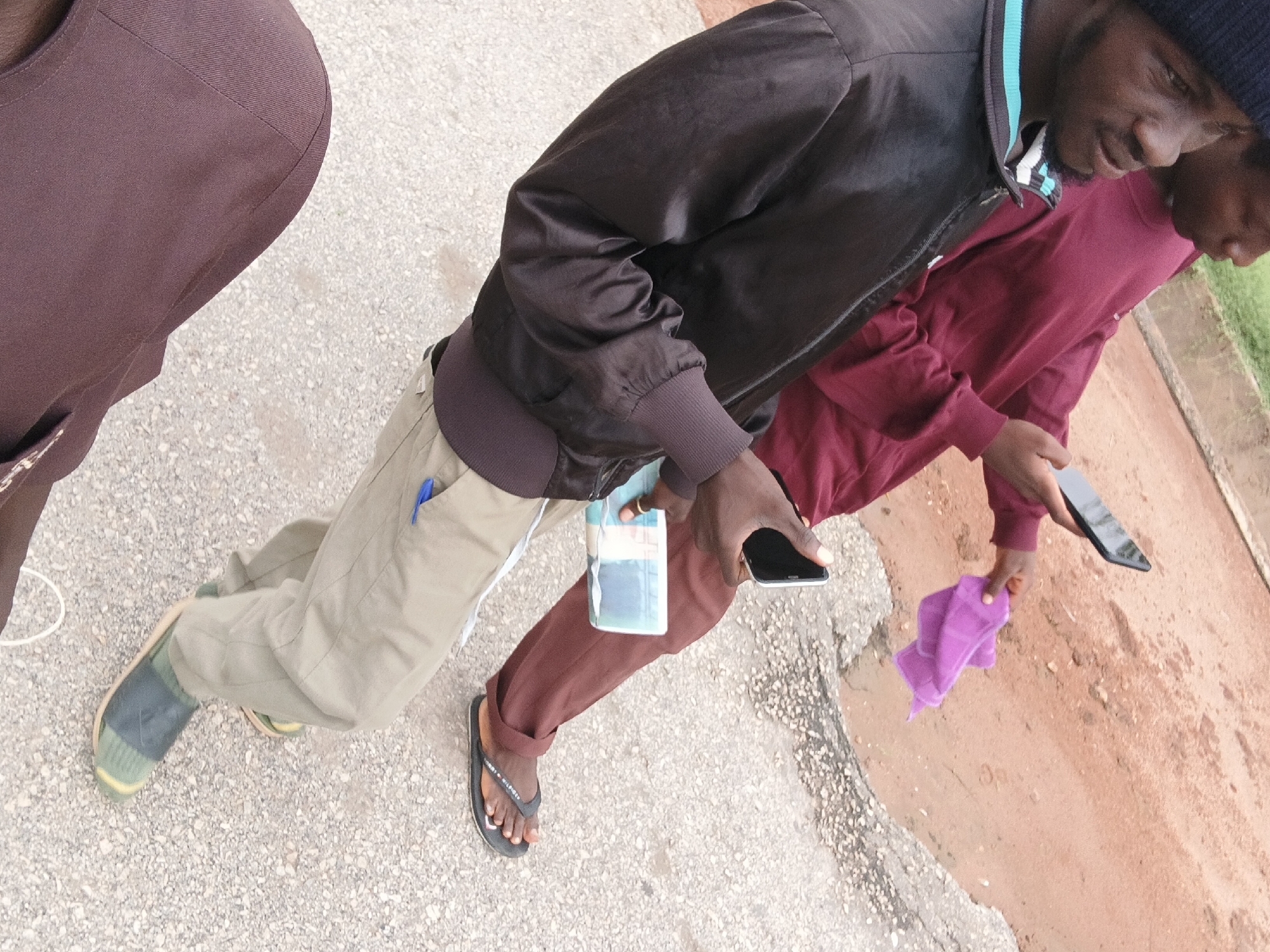 In Jalingo, Nigeria, two individuals are captured in a close-up outdoor shot. The person in the foreground, wearing a black jacket with a blue and white striped collar, is seen from the chest down. They are holding a bundle of cash and a smartphone in their left hand. Their right hand is holding another smartphone, angled downwards. The person is wearing light olive-colored pants and dark open-toed sandals with a dark strap. A gold ring is visible on their left ring finger. Behind them, a second individual is wearing a maroon long-sleeved shirt and is also holding a smartphone. This individual is also holding a small, folded purple cloth in their left hand. The ground is a mix of asphalt and gravel. The background shows a reddish-brown dirt path with some vegetation.