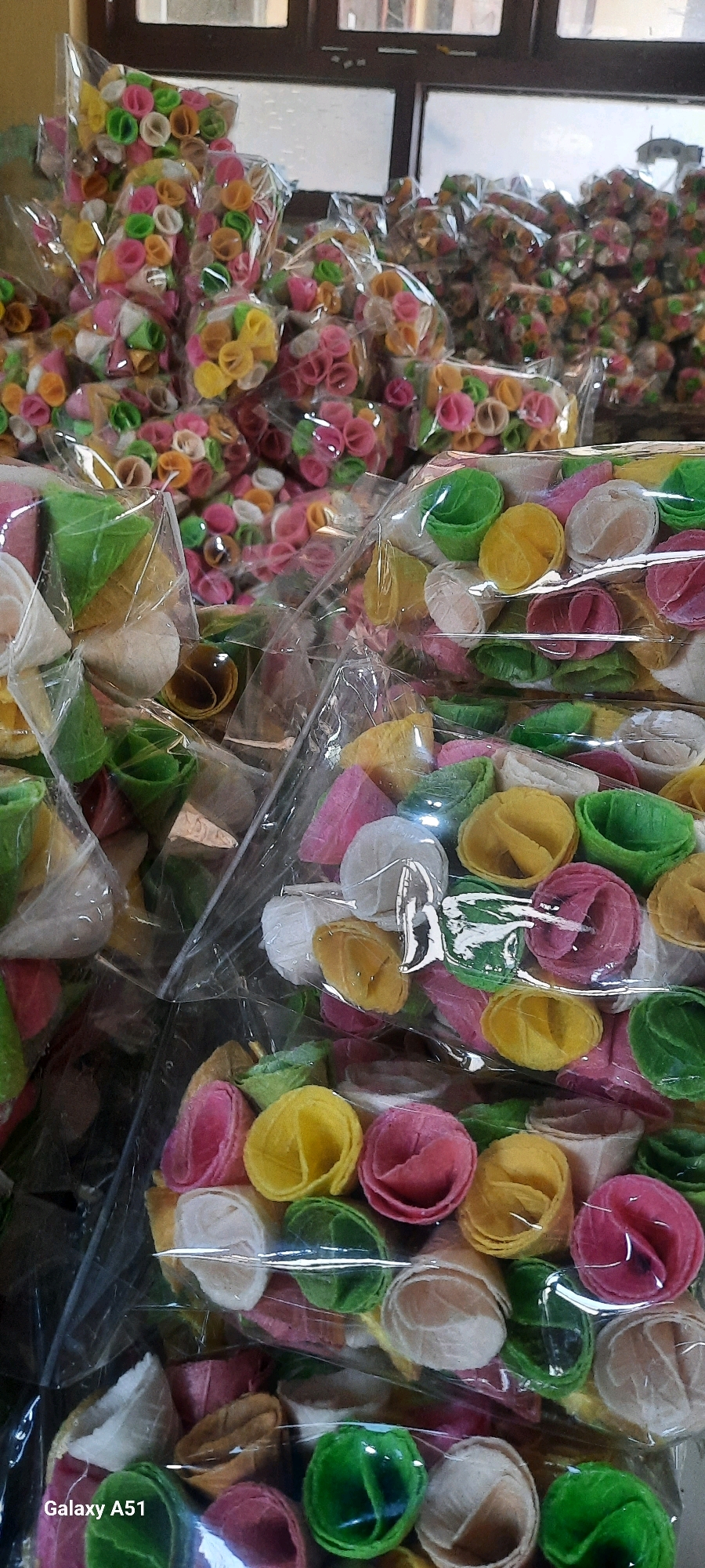 The image shows a close-up of numerous brightly colored, rolled snacks, likely a type of wafer or cracker, packaged in clear plastic. The snacks are predominantly pink, yellow, green, and white, with some appearing to be cone-shaped and others flatter and coiled. They are stacked in multiple bags, some in the foreground and others further back, creating a textured and visually dense composition. The background is blurred but suggests an indoor setting with wooden elements and windows, possibly a market stall or a production area. A "Galaxy A51" watermark is visible in the lower left corner, indicating the device used for capture. The scene appears to be a commercial display of food products.