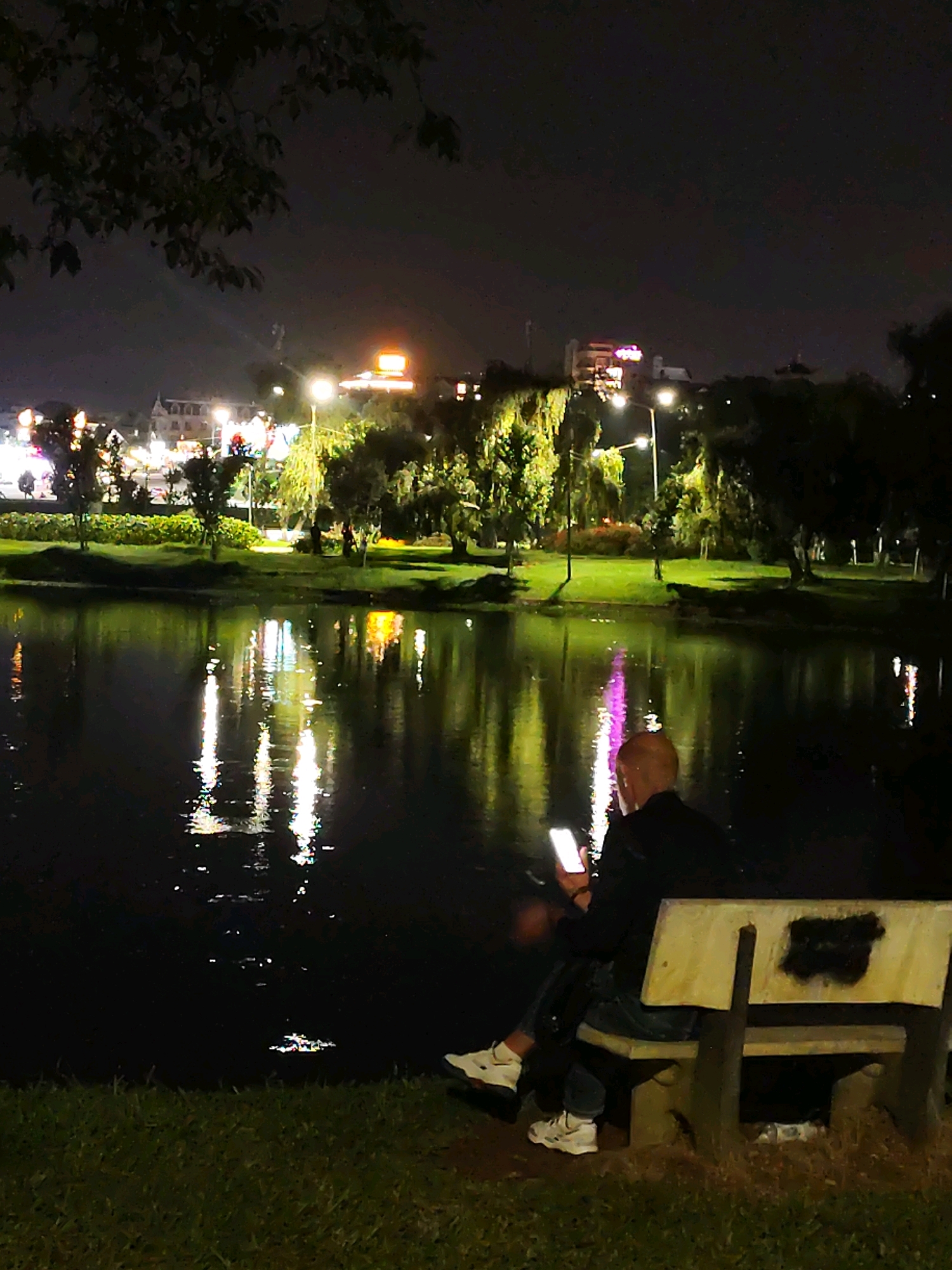 This image depicts a nighttime scene at Xuan Huong Lake in Da Lat, Vietnam. A person, appearing to be an adult male with a shaved head, is seated on a concrete park bench to the right of the frame, facing away from the viewer and towards the lake. He is illuminated by the glow of a smartphone he is holding in his hands, which is positioned in front of him. He is dressed in dark clothing, including a jacket and jeans, and is wearing white sneakers. The bench itself is made of concrete with a wooden slat backrest that has black markings on it. The ground in front of the bench is covered in grass and dry leaves.
The lake occupies the majority of the lower half of the image, reflecting the lights from the city on the far bank. The city lights are a mix of yellow, white, and pink hues, illuminating buildings and trees. Streetlights are visible along the promenade, casting long reflections on the dark water. The far bank of the lake is lined with trees and some buildings, suggesting an urban park setting. The overall atmosphere is tranquil and dimly lit, characteristic of a city park at night.