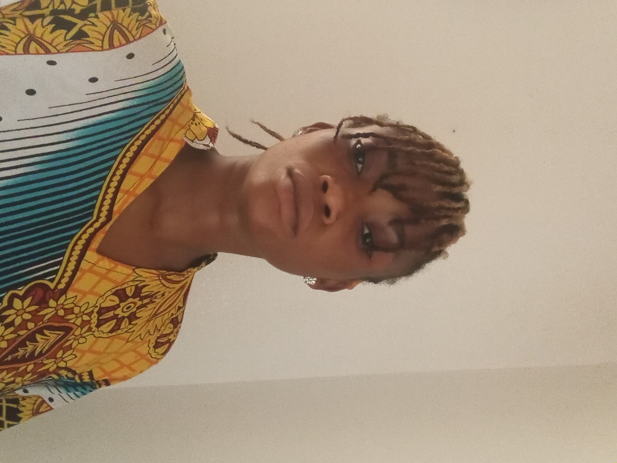 A person with brown braided hair, wearing a patterned yellow, blue, white, and brown garment, is viewed from a low angle against a plain off-white background. The location is Unknown, Unknown.