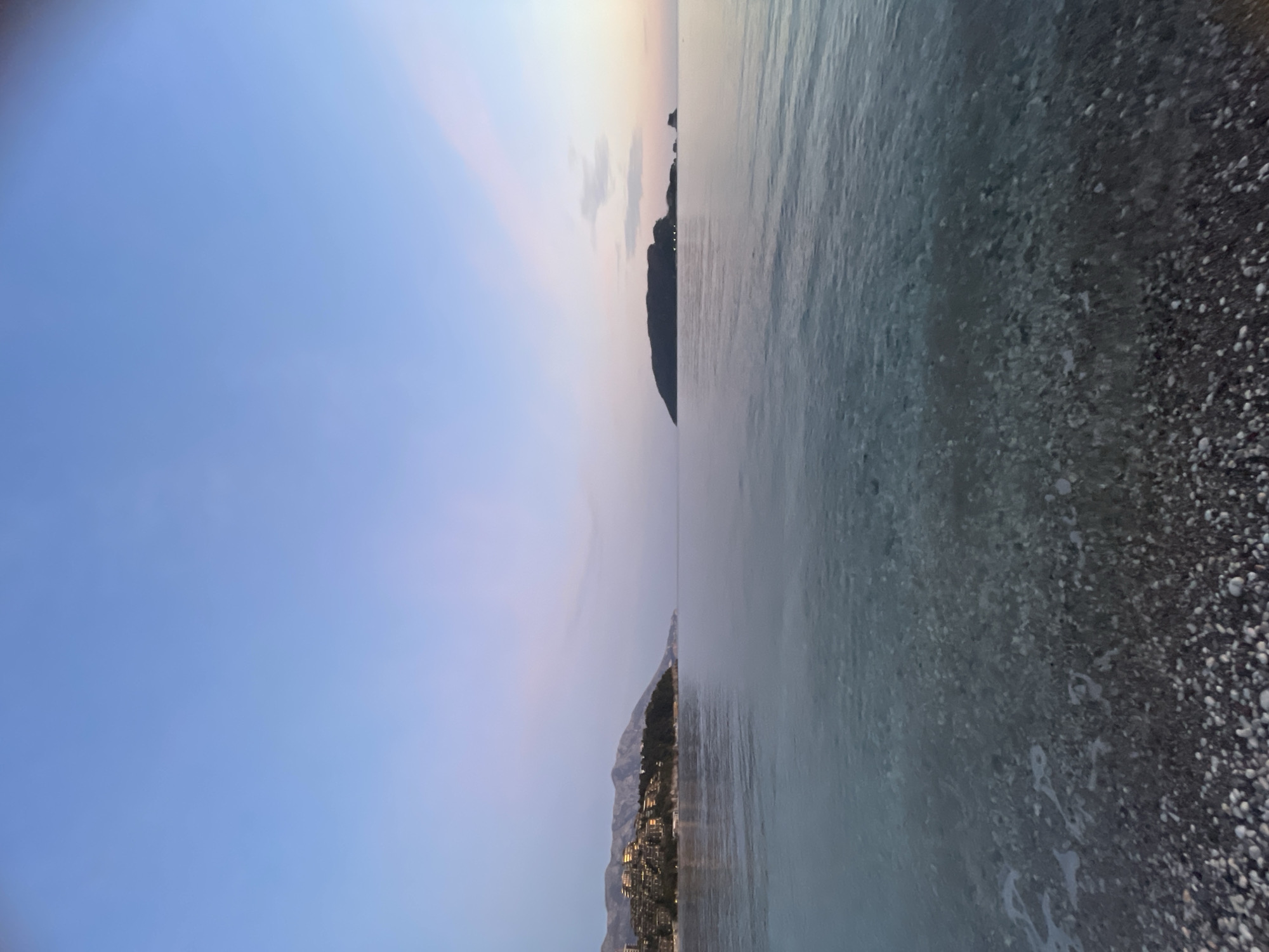 The image depicts a twilight scene from a pebble beach in Budva, Montenegro, looking out over the Adriatic Sea. The immediate foreground shows a dark pebble beach transitioning into shallow, clear water where individual stones are visible beneath the surface. Gentle ripples and minimal foam are present at the water's edge. The midground consists of calm, open sea with a subdued, reflective surface.
To the left, a distant coastline features a series of multi-story buildings, likely resorts or residences, illuminated by interior or exterior lights, situated on a vegetated hillside. Mountains rise in the background behind these structures. To the right, a prominent, dark, irregularly shaped island, identifiable as Sveti Nikola island, is silhouetted against the horizon. Smaller rock formations are visible adjacent to the island, with a few small, bright lights discernible on or near the island itself.
The sky occupies the upper half of the image, displaying a gradient of light blue overhead transitioning to soft pink and lavender hues near the horizon. No people, vessels, or explicit activities are visible. The overall lighting suggests either dawn or dusk.