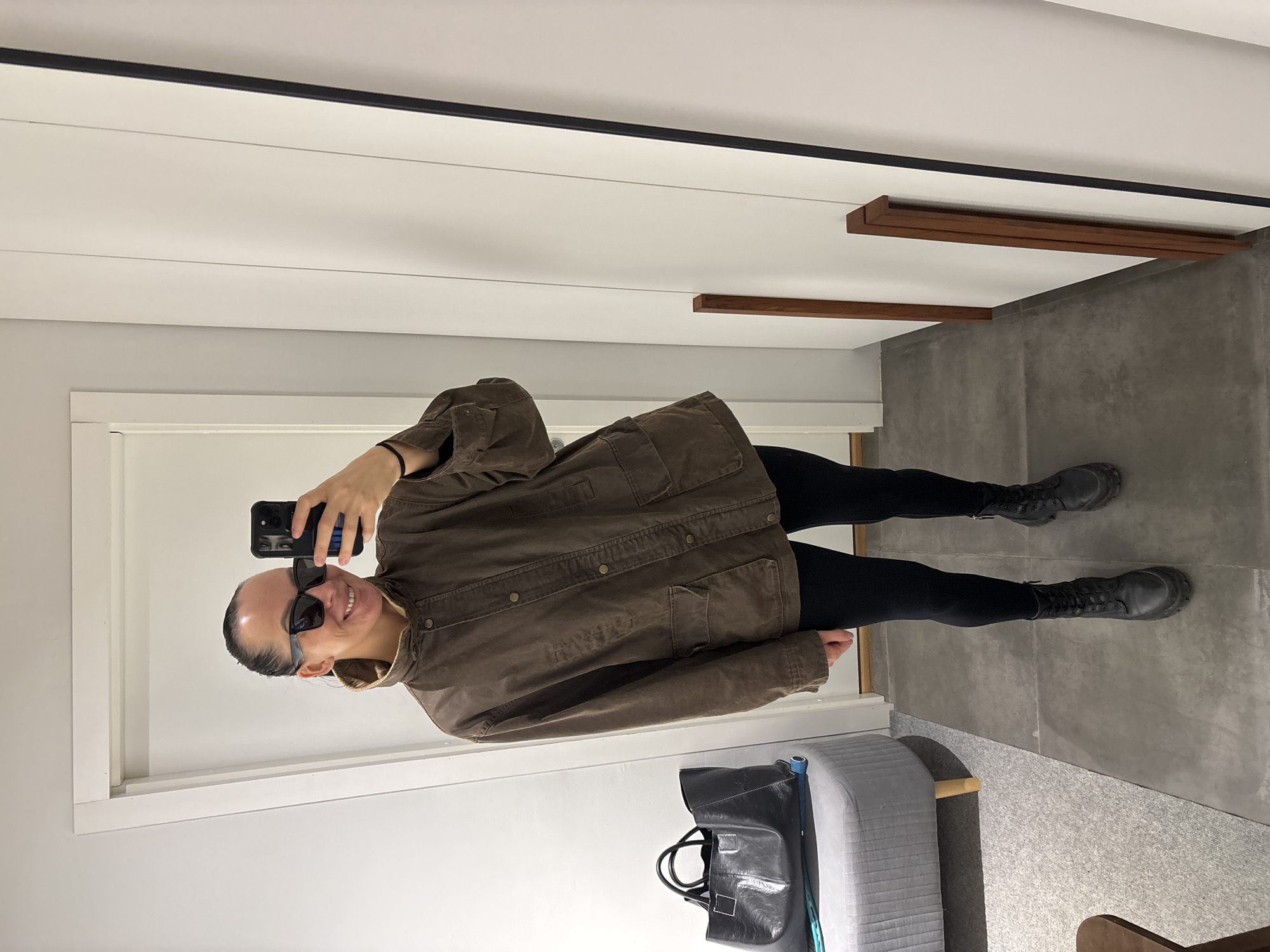 An individual is depicted taking a mirror selfie in an indoor setting in Chaikovsky, Russia. The person, a female-presenting adult, is shown from mid-thigh up, standing on grey tiled flooring. She wears a brown, long-sleeved, collared jacket with multiple buttons down the front and two lower patch pockets. Her attire also includes black leggings and black chunky combat boots with laces and zippers. She has dark hair pulled back, wears black square-framed sunglasses, and smiles towards the camera reflection. Her right hand holds a black smartphone, positioned to capture the image.
To the left of the individual, on a light grey carpeted area, a grey upholstered ottoman or bench is visible. A large black tote bag with white stitching rests on this ottoman, with a blue rolled object partially visible underneath it. A dark brown wooden object is also visible on the carpet in the bottom-left corner. The background features a white doorframe on the left and white walls throughout. On the right, a full-length mirror reflects the scene, flanked by white wall panels and two dark reddish-brown wooden slats leaning vertically against the wall. The environment is well-lit and appears clean and minimalist.
