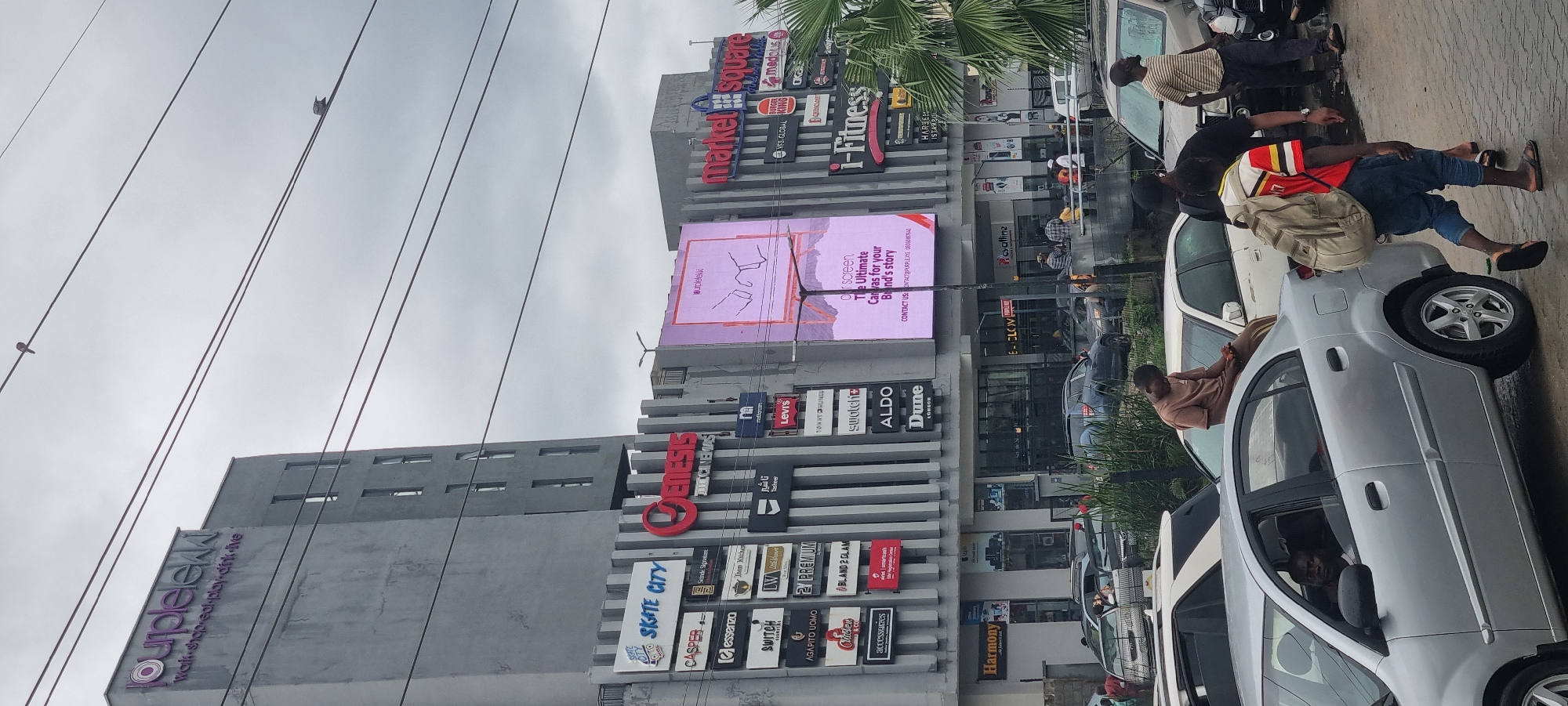 This image captures a street scene in Moba, Nigeria, featuring a multi-level commercial building adorned with numerous signs for various shops and services, including "Market Square," "i-Fitness," "Burger King," "Levi's," "Aldo," and "Harmony." A large digital billboard displays an advertisement with the text "Our screen. The Ultimate Canvas for your Brand's story."
Several people are visible in the scene. Some are standing near parked cars, while others appear to be interacting with the storefronts. One person is leaning against a silver car, and another is sitting in the driver's seat of a silver vehicle. A person in the foreground is carrying a backpack.
The setting is an urban environment with a cloudy sky, suggesting overcast weather. The ground appears wet, possibly from recent rain. The building's architecture is modern, with rows of windows and prominent signage. The overall impression is a busy commercial area, typical of a city in Nigeria. The time of day is not explicitly indicated but appears to be daytime.