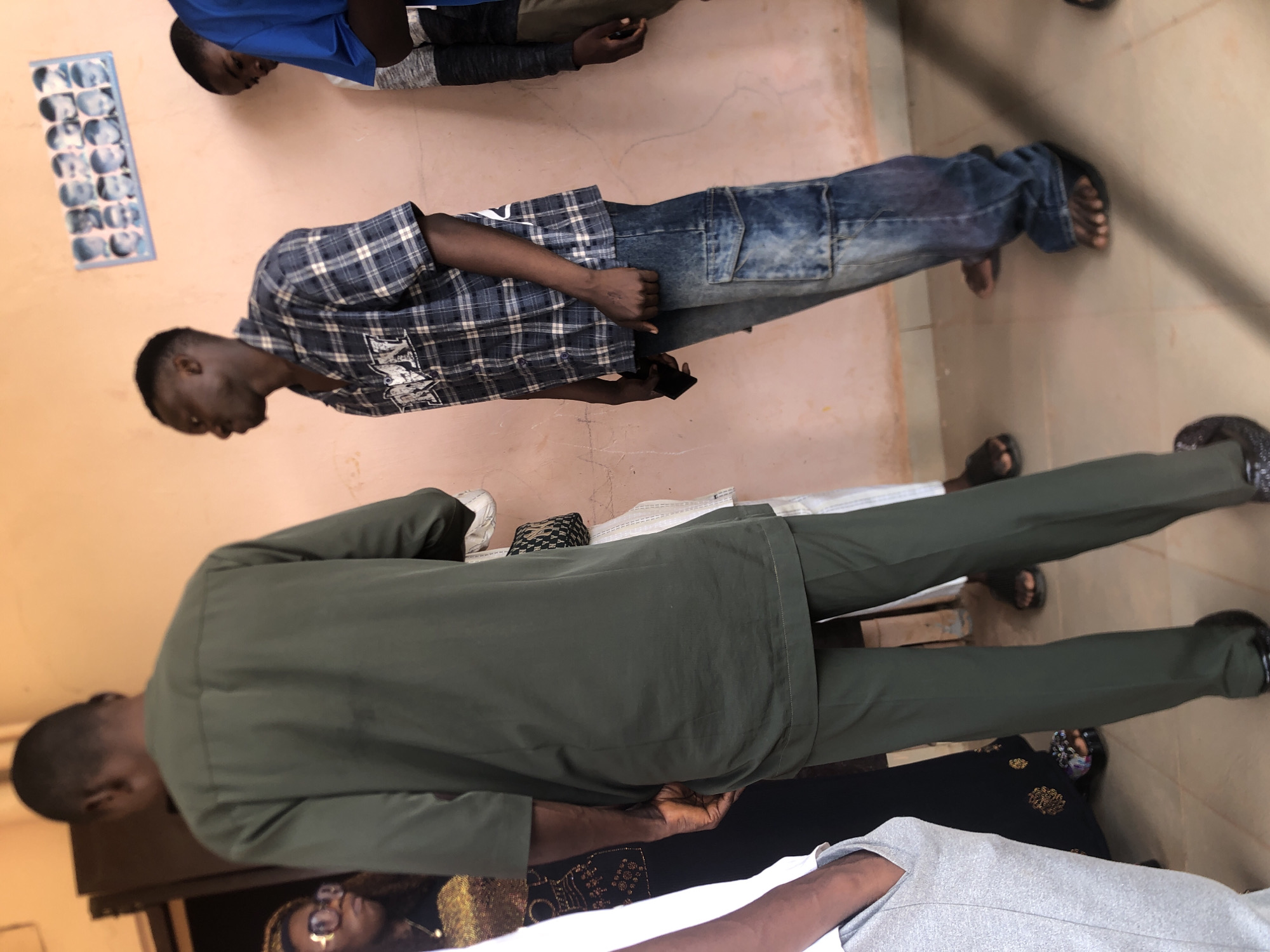 The image shows a group of people standing indoors. In the background, a person in a blue shirt and another in a plaid shirt and jeans are visible. Further in the foreground, a person is wearing an olive green suit with matching pants and black shoes. Additional people in casual attire are partially visible at the bottom of the frame. A wall with a framed set of images hangs in the upper left. The location is Nkwelle, Nigeria.