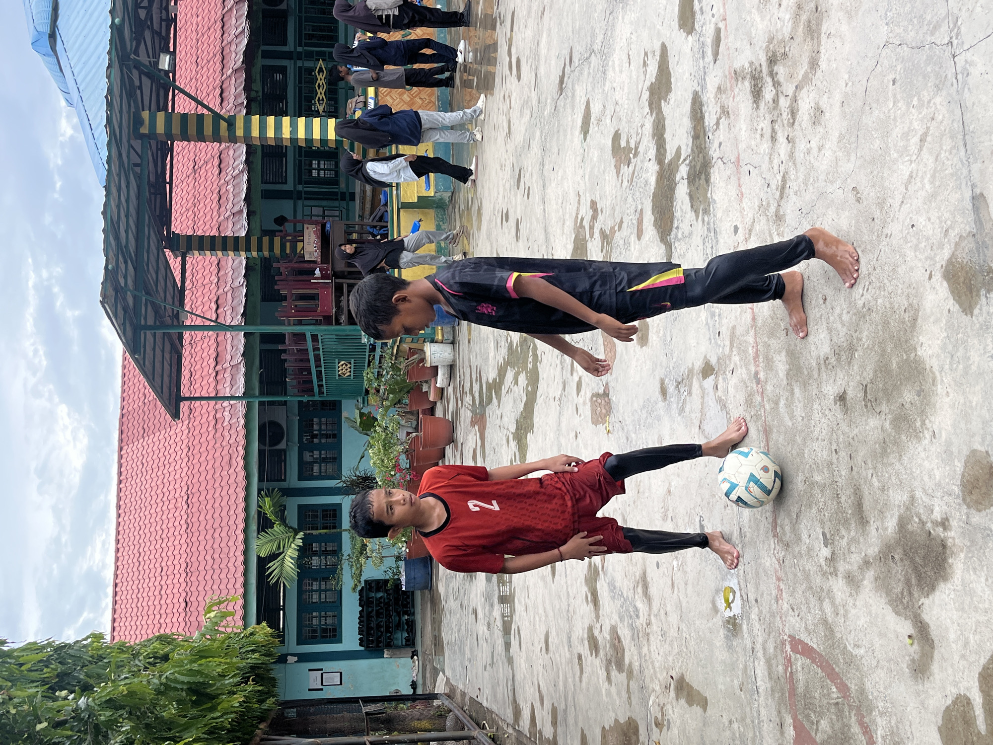 The image depicts an outdoor scene, likely on the grounds of a school in Lhokseumawe, Indonesia, given the architecture and attire of the individuals. Two young boys are the primary subjects, positioned in the foreground on a wet, concrete surface. The boy on the left wears a red jersey with the number '2' prominently displayed, red shorts, and black leggings. He is barefoot. The boy on the right wears a black and pink soccer uniform and is also barefoot. A white and blue soccer ball rests between their feet. In the background, a building with a red tiled roof and light blue walls is visible, with several other individuals, mostly young women wearing hijabs, standing under a covered walkway. The environment appears to be an open courtyard with some trees and potted plants. The ground is wet, suggesting it may have recently rained.