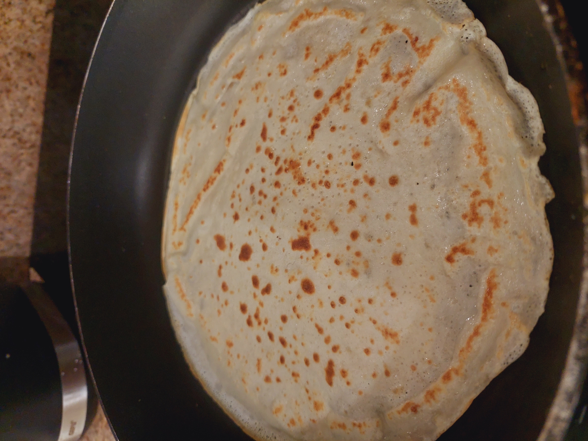 A close-up, overhead shot shows a thin, cooked crepe with golden-brown speckles and browned edges, resting in a black non-stick frying pan. The crepe's surface is slightly textured and glistening, indicating moisture or oil. The pan is situated on a speckled brown countertop. In the upper left background, a black kitchen appliance with a silver band is partially visible. The scene is an indoor domestic setting, likely a kitchen. There are no people visible in the image. The crepe is likely being cooked or has just been cooked. The image does not provide any information that would allow for localization to Sofia, Bulgaria.