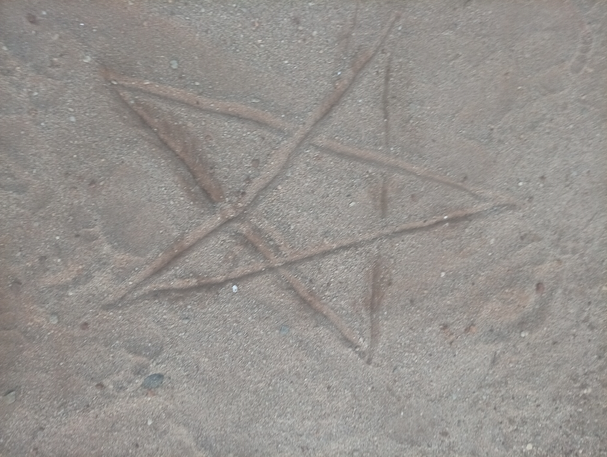 The image depicts a pentagram drawn in sand. The sand appears coarse and light brown in color. The pentagram lines are created by impressions in the sand. The location is unknown, and there is no information to indicate it is in Ikorodu, Nigeria. No people or other objects are visible. There is no action taking place.
