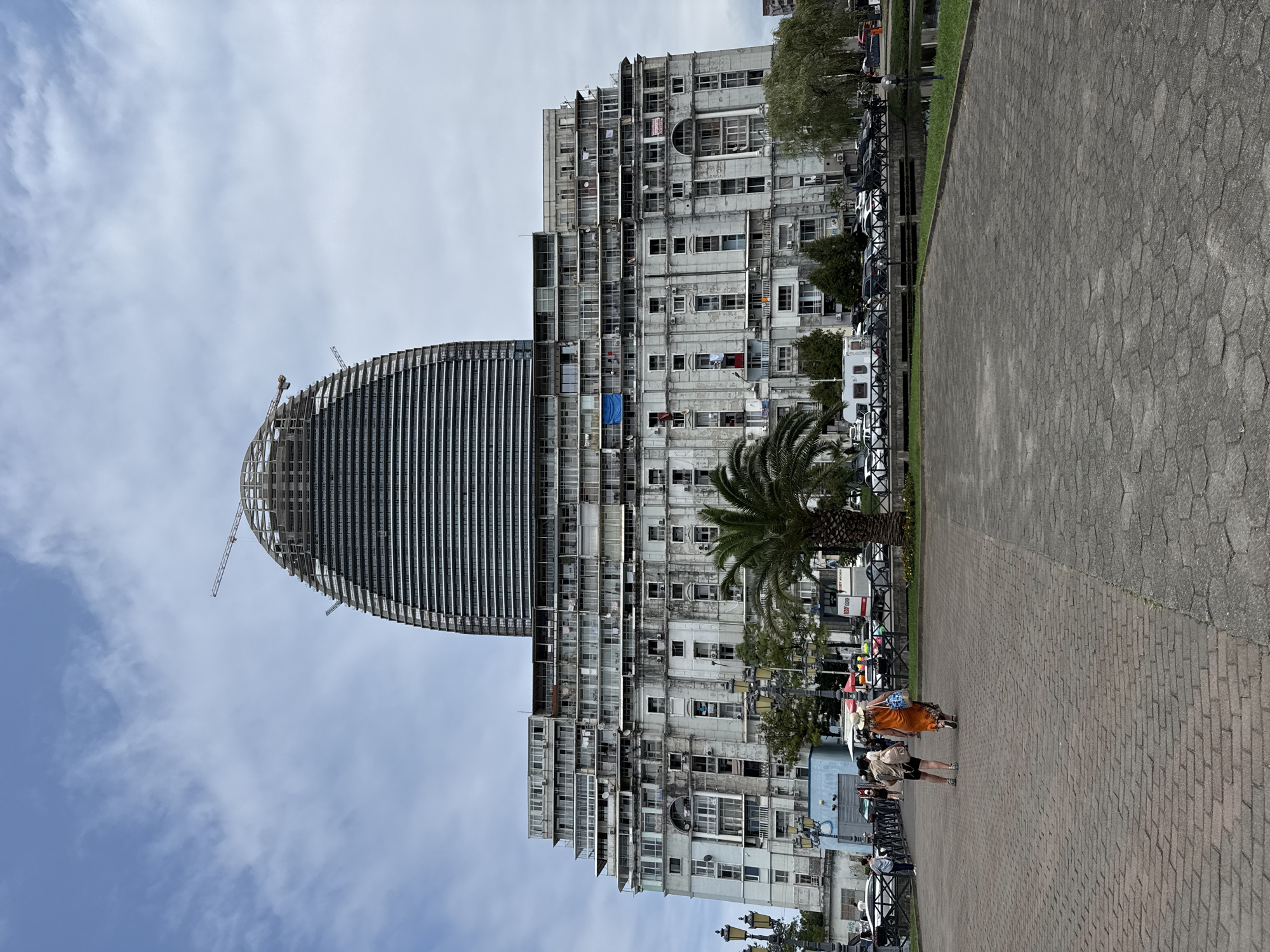 A wide shot shows a massive, partly constructed building with a distinctive curved top resembling an eggshell, possibly under renovation or extension, in an urban setting. The building's lower section is a multi-story concrete structure with numerous balconies, many displaying air conditioning units and laundry hanging out to dry. Two individuals are walking on a brick-paved walkway in the foreground. To the left of the walkway are streetlights and to the right, a palm tree, a body of water, and parked cars. The sky above is cloudy. The scene is an outdoor view of a city.