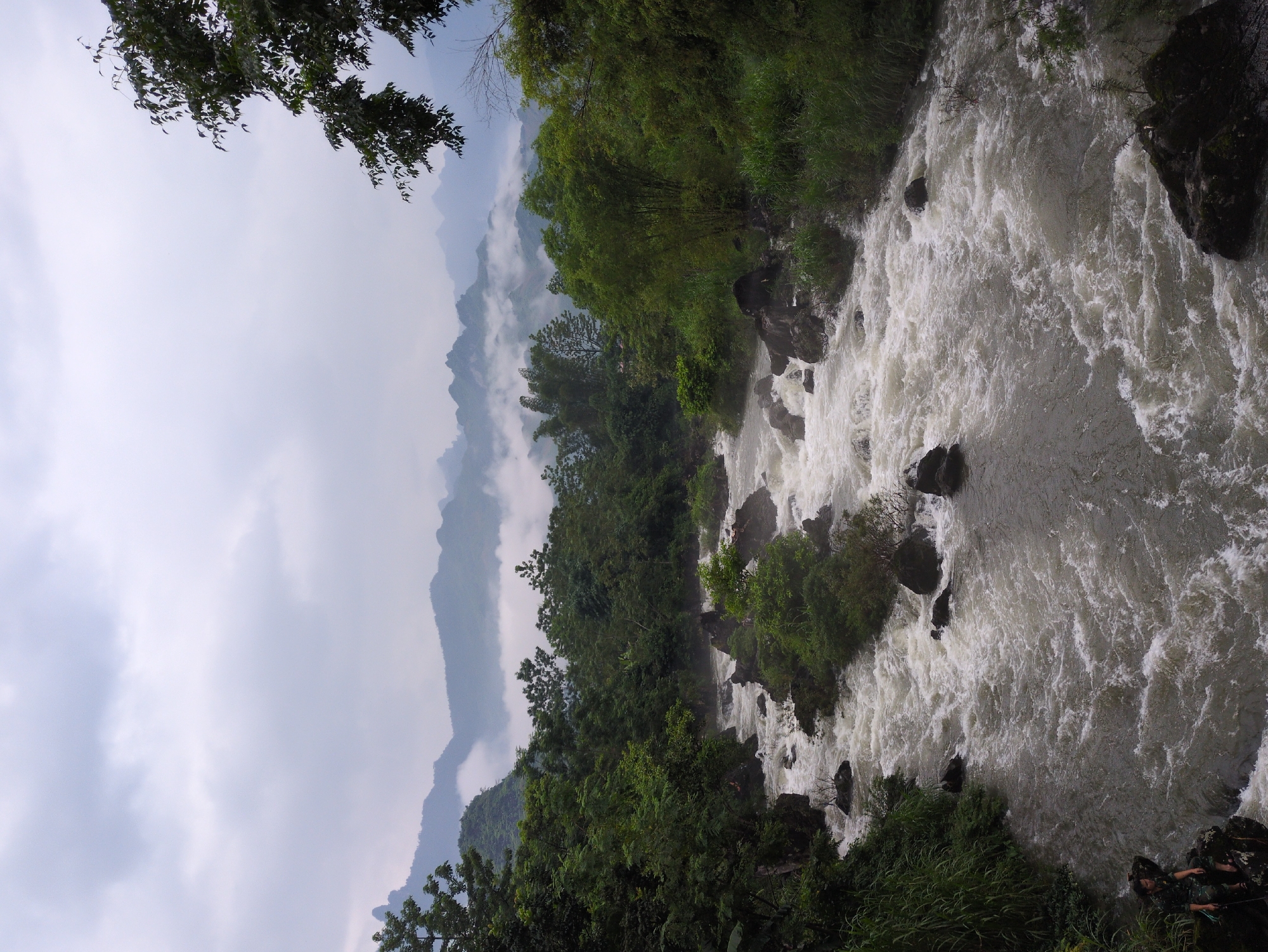 A wide, turbulent river flows through a densely vegetated valley under a cloudy sky. Mountains are visible in the background, shrouded in mist. The river is characterized by white water rapids and numerous dark rocks protruding from the churning current. Lush green trees and foliage line the banks of the river, with some vegetation also growing on the rocks within the water. In the foreground on the left bank, a person in camouflage clothing is sitting on a dark rock, looking towards the river. The overall scene depicts a natural, somewhat wild landscape with a focus on the powerful movement of water. This scene is located in the Du Già Commune, Vietnam, a region known for its natural beauty and mountainous terrain.