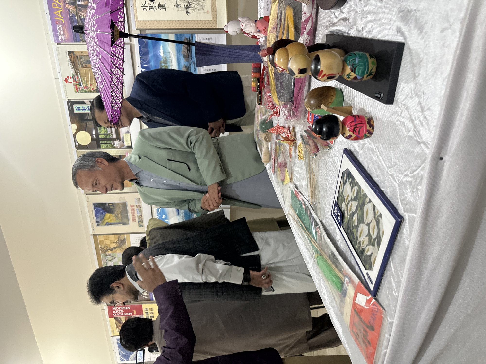 The image shows a group of people gathered around a table displaying various objects. The table is covered with a white tablecloth and holds items such as wooden dolls, origami cranes, and framed artwork. People are standing around the table, looking at the items. A man in a light green suit stands near the table, his hands clasped in front of him. A purple umbrella is visible in the background. Posters and calendars with Japanese writing and imagery hang on the wall. The environment suggests an art exhibit or cultural display. Based on the OCR results, there are calendars from "Highway in Japan" and signs with the names "Modern Arts Gallery" and Japanese text. These details suggest the image was taken at a Japanese art exhibition. It is impossible to identify the location in the city of Quetta City Tehsil, Pakistan based solely on the provided image.