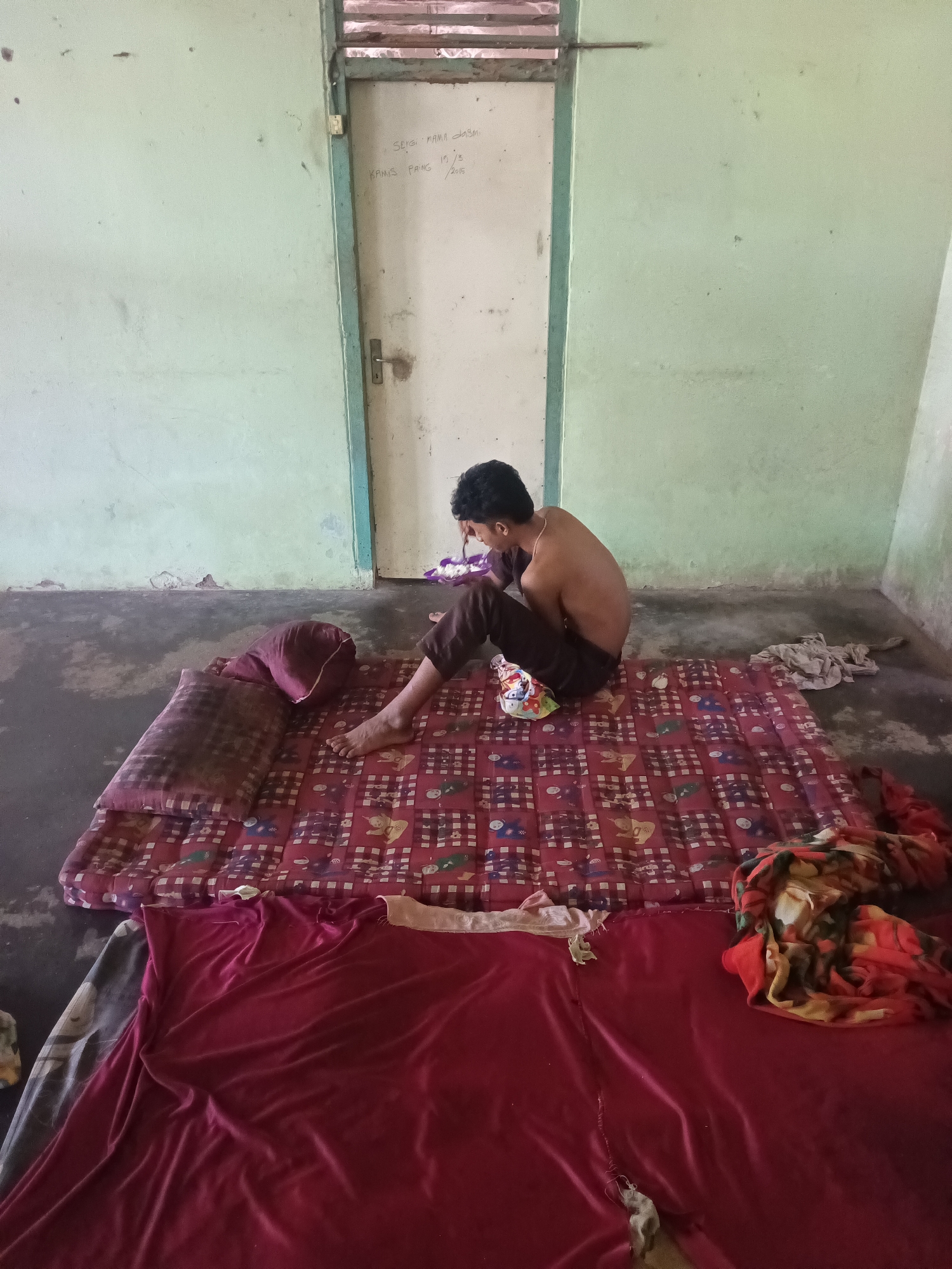 In a sparsely furnished room with pale green walls and a concrete floor, a young man sits shirtless on a red and white checkered mattress. He is wearing dark brown pants and is hunched over, appearing to be looking down at something in his hands. Two maroon pillows are propped up on the mattress beside him. To the right of the mattress, a pile of colorful fabrics lies on the floor. A white door with a simple metal handle is visible in the background, with faint writing in black marker on its surface. The overall impression is one of a simple, perhaps temporary, living space. The writing on the door, as can be seen from a close-up crop, says "SENGI MAMA JASMI" and "KAMIS PAING 15/3/2015". This writing suggests the location is within the Terbanggi Marga area of Indonesia, given the date format and common Indonesian naming conventions.