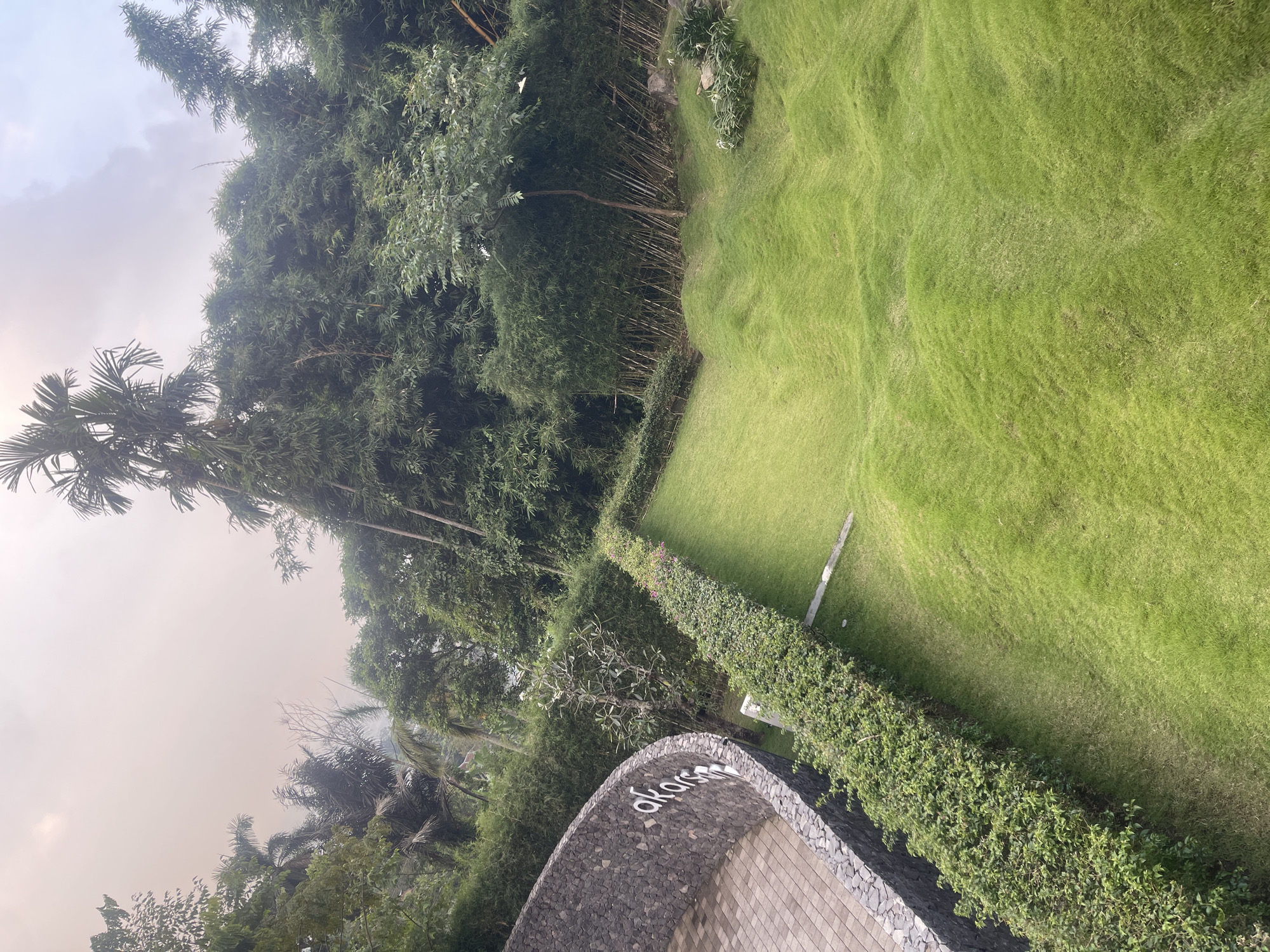 In Pecalukan, Indonesia, an outdoor scene unfolds with lush green grass forming undulating mounds that descend into a lower area. A manicured hedge runs along a curved, stone-clad wall, which is paved with interlocking bricks. The stone wall features the word "akars" in white lettering. Beyond the hedge, a dense bamboo forest stands tall, interspersed with palm trees and other tropical vegetation. The sky above is a pale, hazy orange, suggesting either dawn, dusk, or a smoky atmosphere.