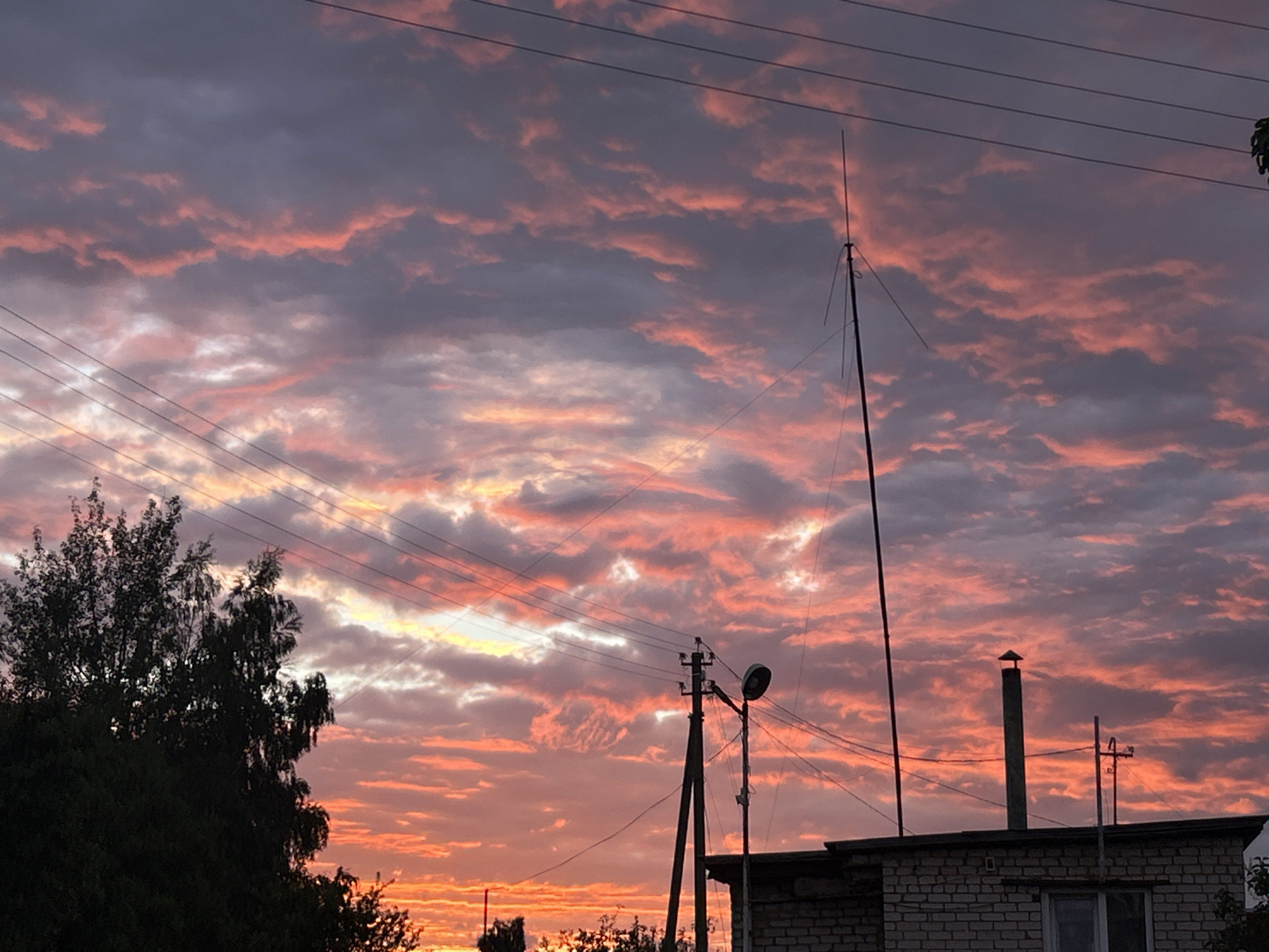 The image captures a dramatic sunset over a rural landscape in Забалоцкі сельскі Савет, Belarus. The sky is ablaze with vibrant hues of orange, pink, and purple, with clouds casting long shadows across the scene. Silhouetted against the colorful sky are several utility poles with wires, a tall antenna or lightning rod, a single-story brick building with a chimney, and a dense tree on the left side of the frame. The overall impression is one of natural beauty and rustic charm.