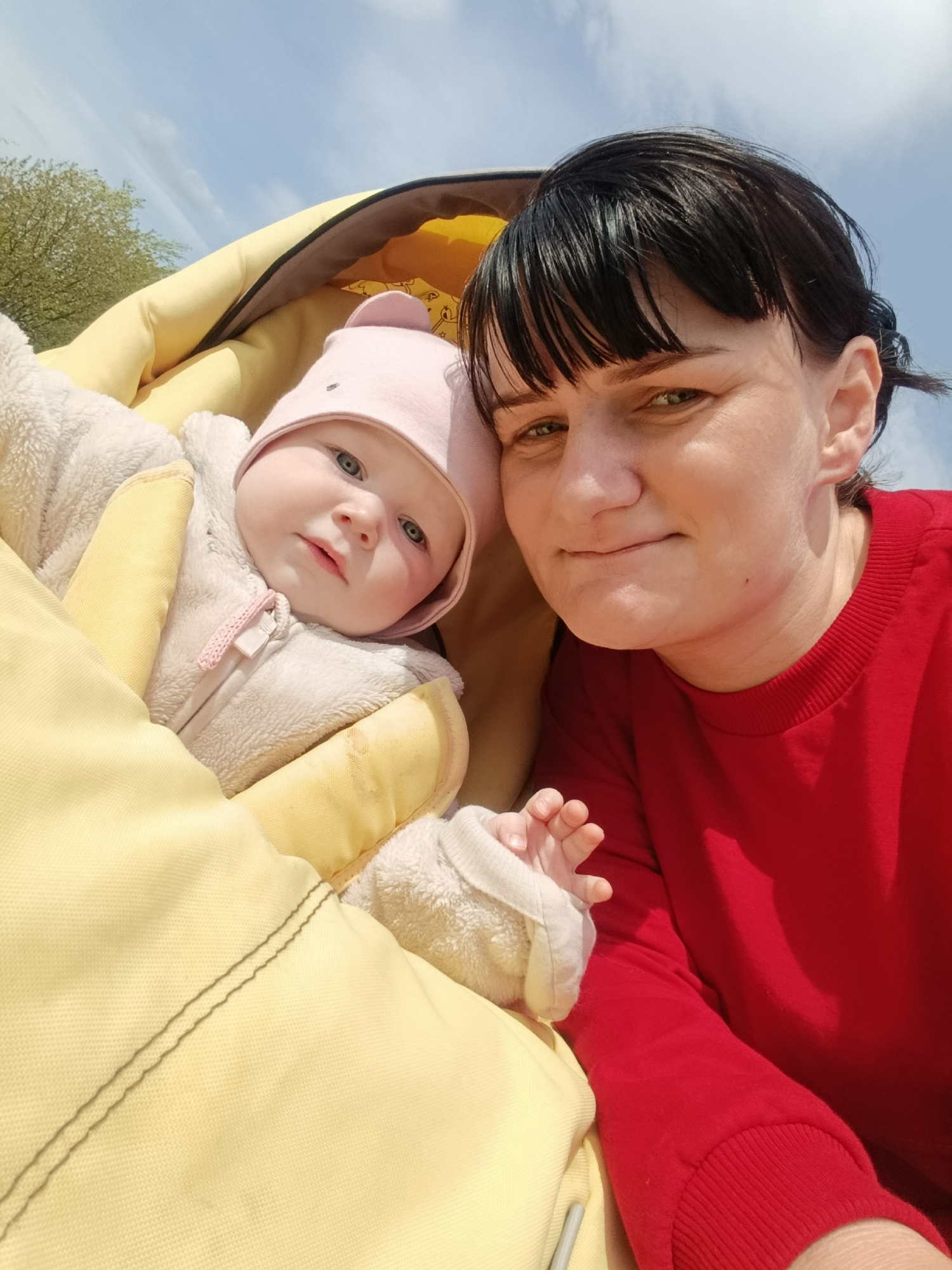 A medium shot depicts a woman and an infant in a stroller on a sunny day, seemingly in Фоки, Russia. The woman, positioned to the right, wears a red long-sleeved shirt and has short, dark hair with bangs. She looks directly into the camera with a slight smile. To her left, an infant lies in a yellow stroller or carrier, bundled in a white, fleece-like suit and a pink hat. The baby's right hand is visible, extending towards the woman. The stroller's interior lining appears to be yellow with a brown trim. In the background, hints of trees are visible on the left, with a blue sky above.