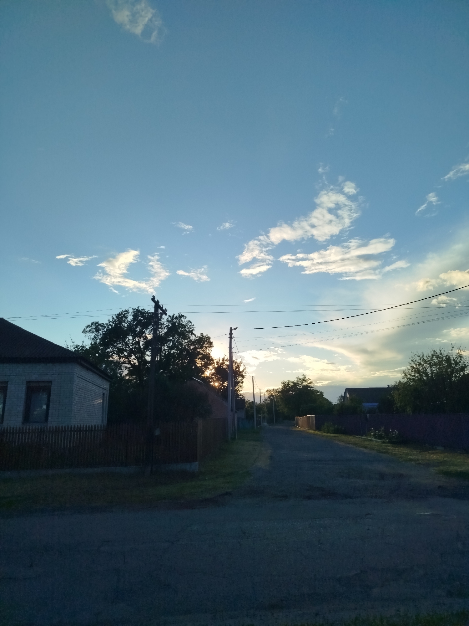The image depicts a rural street scene in Ustyvytsia, Ukraine. The sky is predominantly blue with scattered cumulus clouds, some tinged with a golden hue from the setting sun. A dirt and asphalt road stretches into the distance, with a slight curve. On the left side of the road, there is a small, single-story brick house with a dark roof and a wooden fence. Utility poles with power lines run along the side of the road. Vegetation, including trees and bushes, is present on both sides of the street. The sun is setting in the distance, partially obscured by trees, creating a silhouette effect. The overall lighting suggests it is late afternoon or early evening. No people or animals are visible.