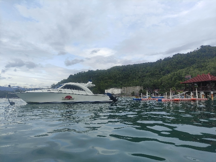 The image presents a tranquil coastal scene in Watulimo, Indonesia, under an overcast sky. The foreground is dominated by calm, clear water exhibiting gentle ripples, hinting at a placid bay or inlet.
A sleek, white speed boat, or yacht, is moored or anchored slightly to the left of the center. Its hull features red and black lettering that clearly reads "Lady Say," with additional, smaller black text above it that is difficult to discern, possibly a registration number. A small, dark anchor buoy or marker is visible in the water near the bow of the boat.
Towards the right, closer to the shore, a series of colorful floating platforms and buoys, in shades of blue, pink, and orange, are arranged on the water, suggesting a recreational area or a resort's dock. Behind these floats, a wooden structure with a distinctive red, tiered roof stands on stilts over the water, resembling a gazebo or a waterfront building.
The background is characterized by a dense, verdant hillside covered in lush tropical trees, rising steeply from the water's edge. A light-colored, possibly concrete, building with a flat roof can be seen partially obscured by the foliage at the base of the hill. The sky is overcast with soft, diffused light, indicating daytime, possibly late morning or afternoon. No people are visible, contributing to the serene and untouched atmosphere of the scene. The overall impression is one of a peaceful tropical getaway focused on water activities and natural beauty.