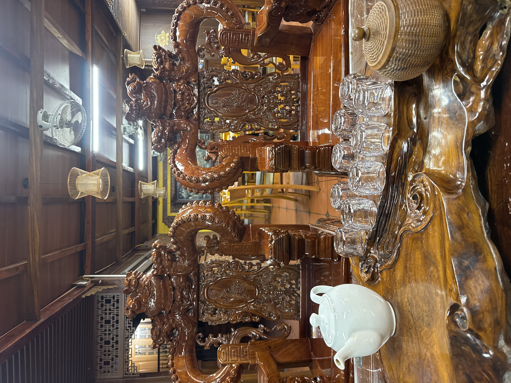 The image depicts a traditional Vietnamese tea setting, likely within a restaurant or home in Mộc Châu, Vietnam. Two intricately carved wooden chairs, featuring dragon motifs, dominate the foreground. In front of the chairs is a polished wooden table with a natural, wavy edge. On the table, a white ceramic teapot sits to the left, accompanied by a row of clear glass tea cups. To the right, a woven wooden container with a lid is present. The background reveals more of the establishment's interior, with wooden beams, ceiling fans, and lighting fixtures suggesting a rustic yet elegant ambiance. The overall scene conveys a sense of cultural heritage and tranquility.