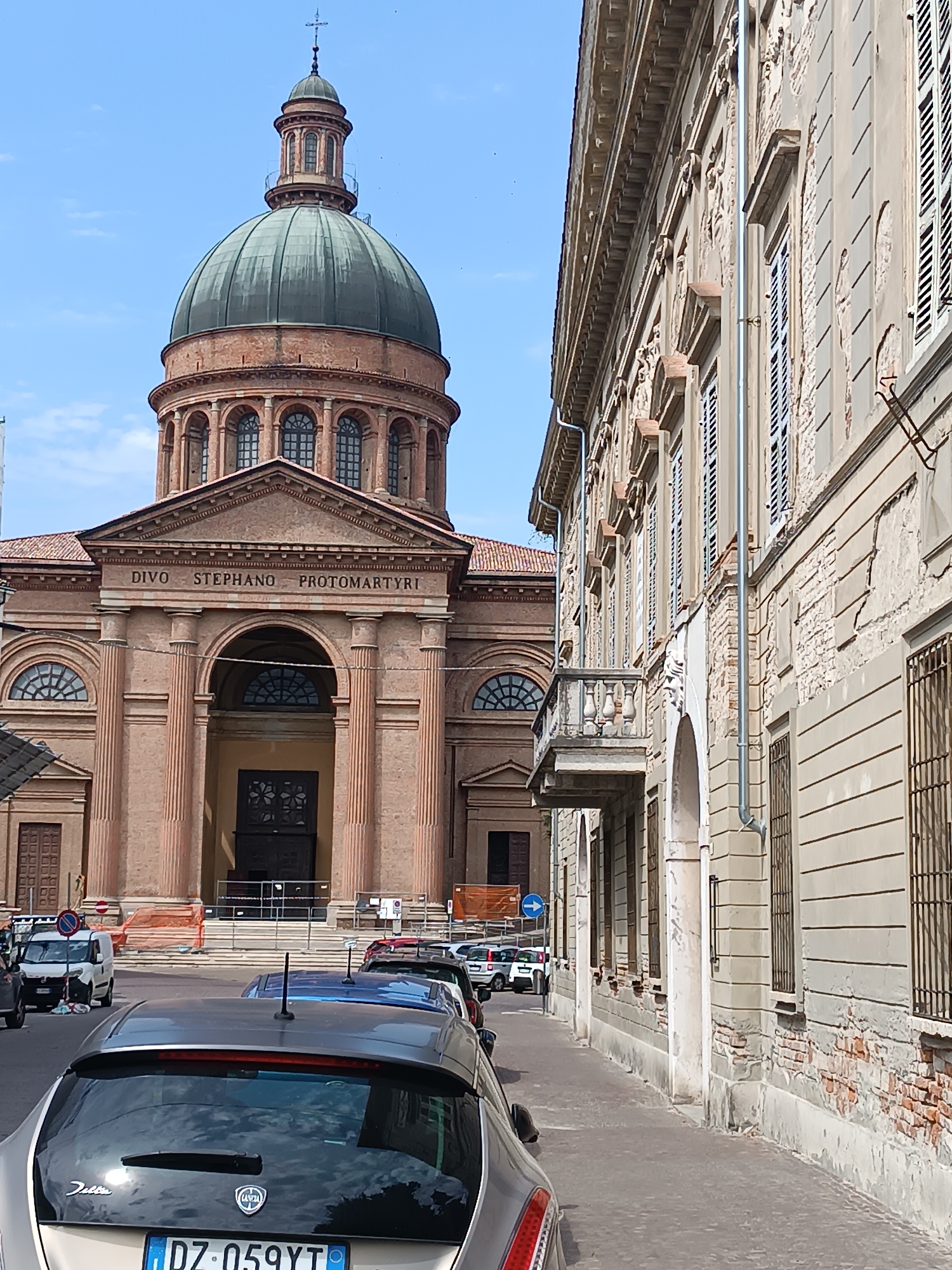 The image shows a street scene in Casalmaggiore, Italy, featuring a large domed building in the background and an older building with peeling stucco on the right. The domed building, identified by the inscription "DIVO STEPHANO PROTOMARTYRI," is likely the Duomo di Santo Stefano, Protomartire, a significant religious structure. In the foreground, several cars are parked on the street. The sky is blue with some scattered clouds. The overall atmosphere suggests a bright, sunny day in an Italian city.