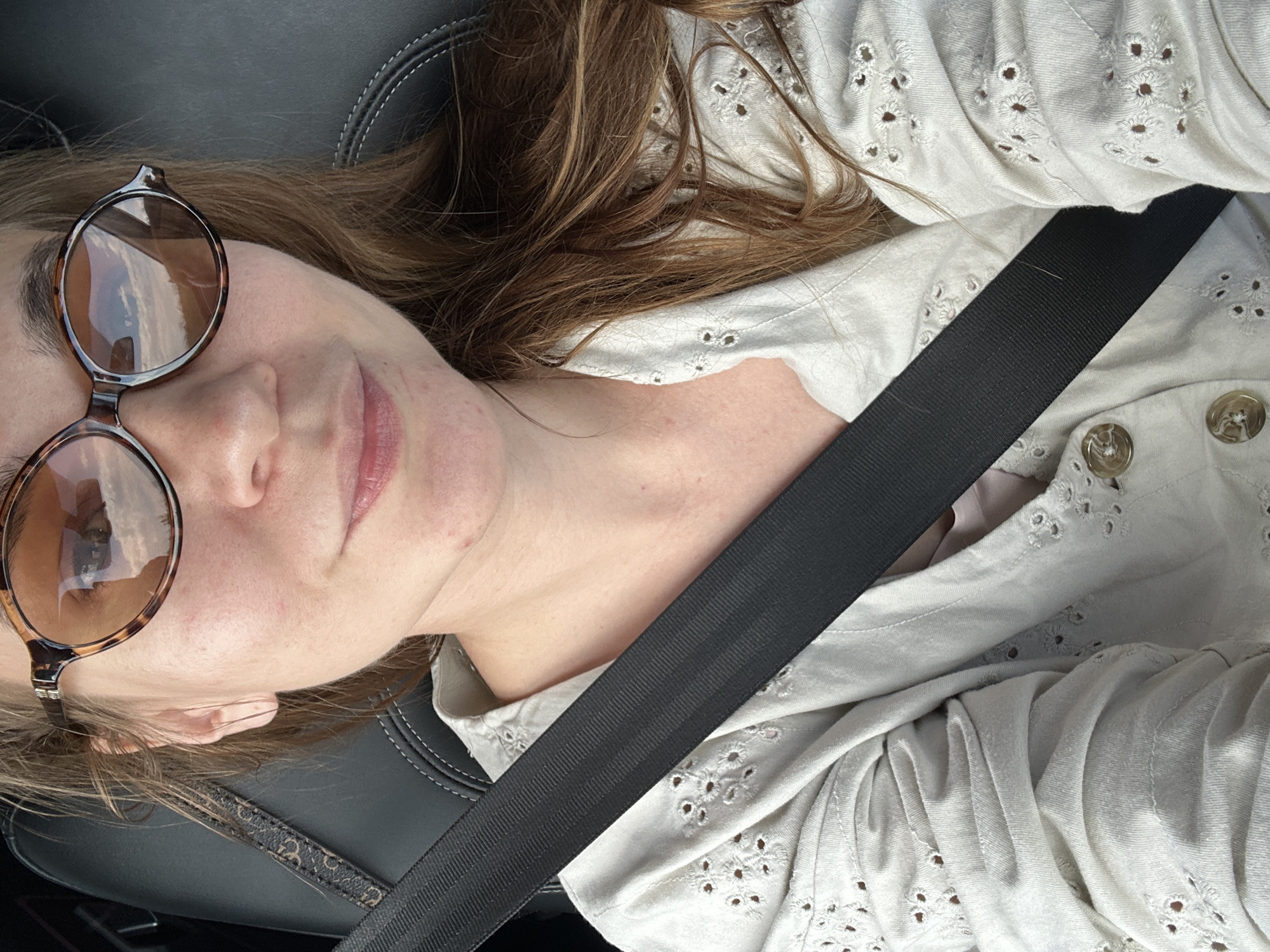 The image shows a close-up, selfie-style shot of a woman wearing tortoiseshell sunglasses and a white eyelet shirt. She is seated in a car, with a black seatbelt diagonally crossing her chest. The background includes the car's dark interior, specifically the headrest and stitching of the seat. The woman's hair is brown and appears to be pulled back loosely. Her expression is relaxed, with a slight smile. The reflection in her sunglasses shows a cloudy sky and what appears to be a landscape. The image does not provide any information or context to place it in the city of Pervomaiskoe, Russia.