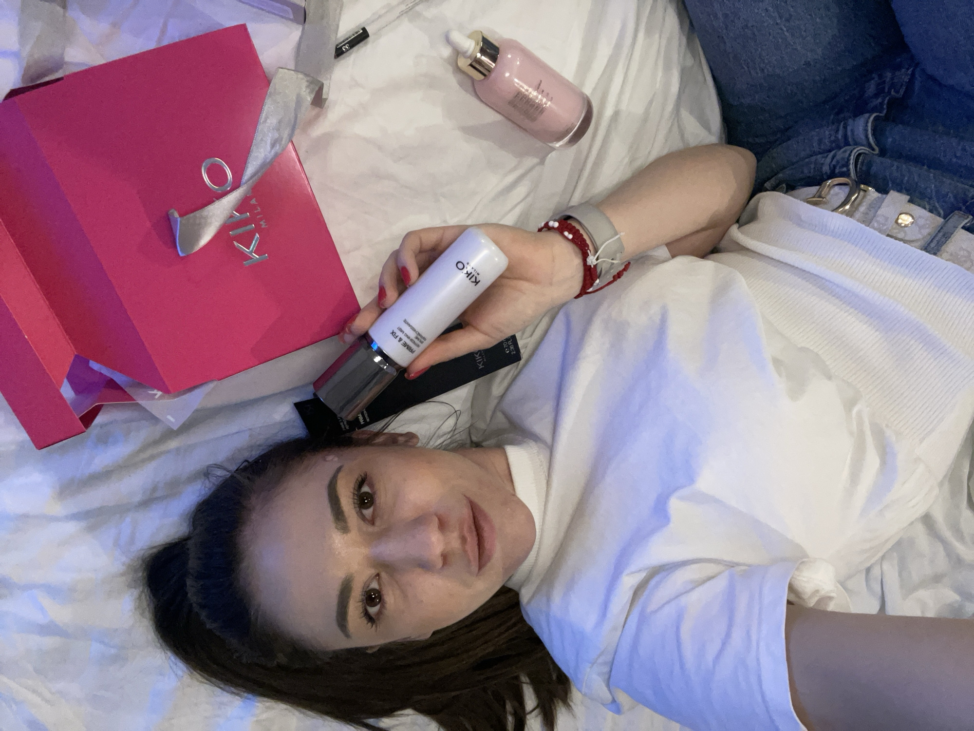 A female individual is depicted lying on a white, rumpled bed surface, viewed from a high-angle perspective, in an indoor setting within Sofia, Bulgaria. She has dark hair and dark eyes, and looks directly towards the camera with a faint smile. She wears a white long-sleeved top and blue denim jeans, secured by a light-colored belt with a gold-toned buckle. Her left hand, adorned with red painted fingernails and multiple wristbands including red and white beaded bracelets and a grey band, holds a white KIKO MILANO "Prime & Fix Refreshing Mist" spray bottle upright. A pink KIKO MILANO "Hydragenia Perfect Glow" dropper bottle lies horizontally on the bed surface below her left hand. To her right, a partially open bright pink KIKO MILANO box with silver lettering and a silver ribbon is present. Additional black KIKO MILANO product packaging is visible near her head and left arm. A small black object marked "33" is situated near the pink box.