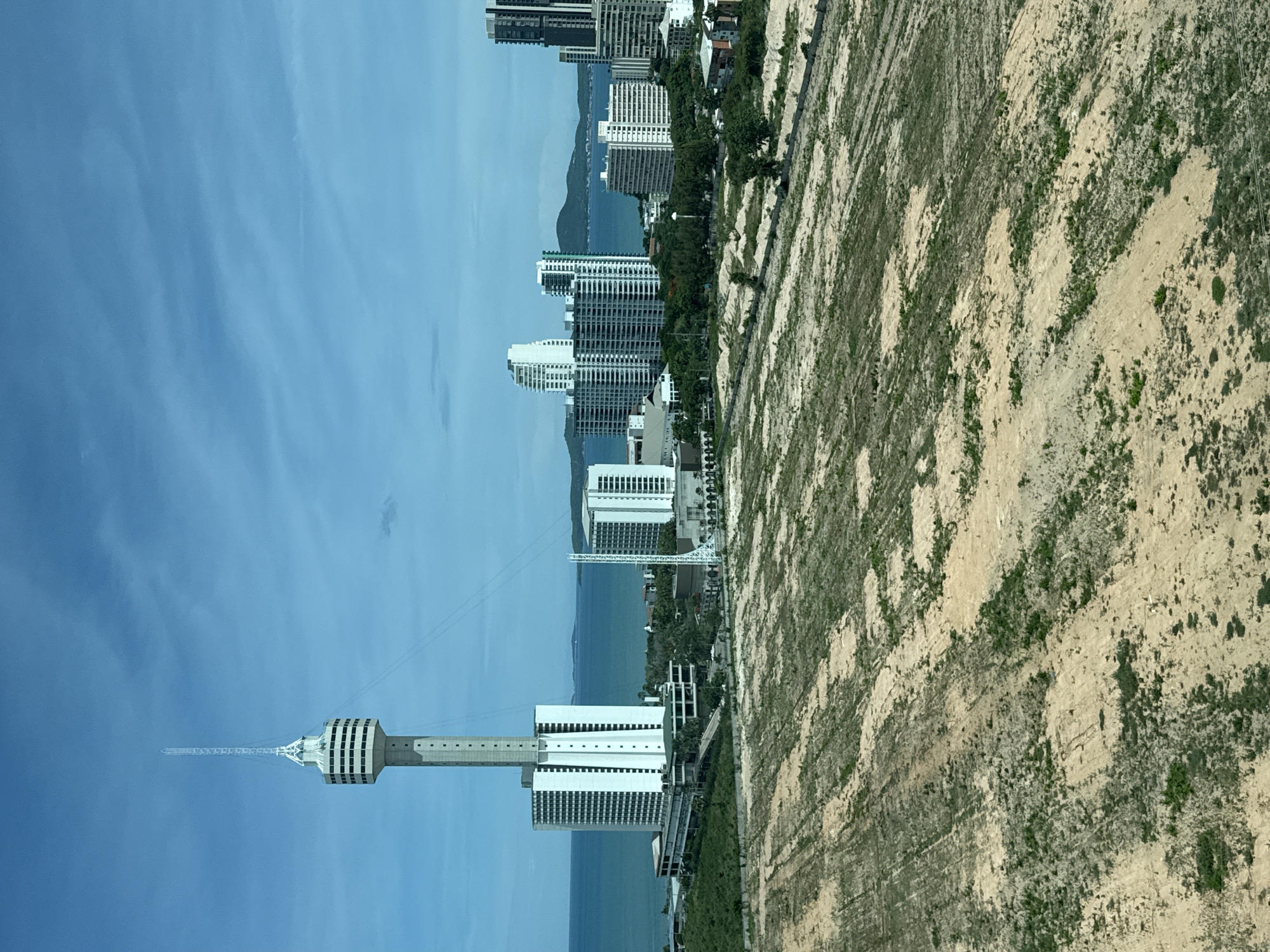 The image shows a panoramic view of Pattaya City, Thailand, with a prominent television tower dominating the skyline. The foreground features a large, undeveloped plot of land with sparse vegetation and visible soil disturbance, suggesting it is a construction site or a recently cleared area. In the mid-ground, several modern high-rise buildings, likely hotels or residential apartments, are visible along the coastline. The blue ocean stretches out beyond the city, with distant islands or landmasses on the horizon. The sky is a clear blue with some wispy clouds. The overall scene depicts a developing urban landscape adjacent to a coastal environment.
