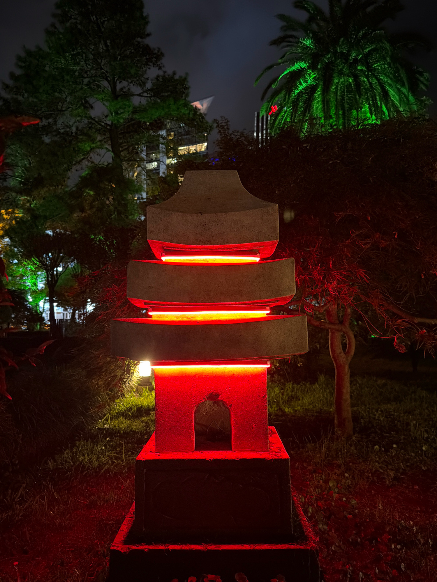 In Batumi, Georgia, at night, a multi-tiered stone pagoda-style lamp stands in the foreground, illuminated by vibrant red neon lights within its segments. The lamp is situated on a dark base, surrounded by grass and a sparse tree with twisted branches on the right. In the background, a building with illuminated windows is visible through trees. To the far right, a palm tree is brightly lit with green lights. The overall atmosphere is moody and atmospheric, with dark shadows and artificial lighting dominating the scene.